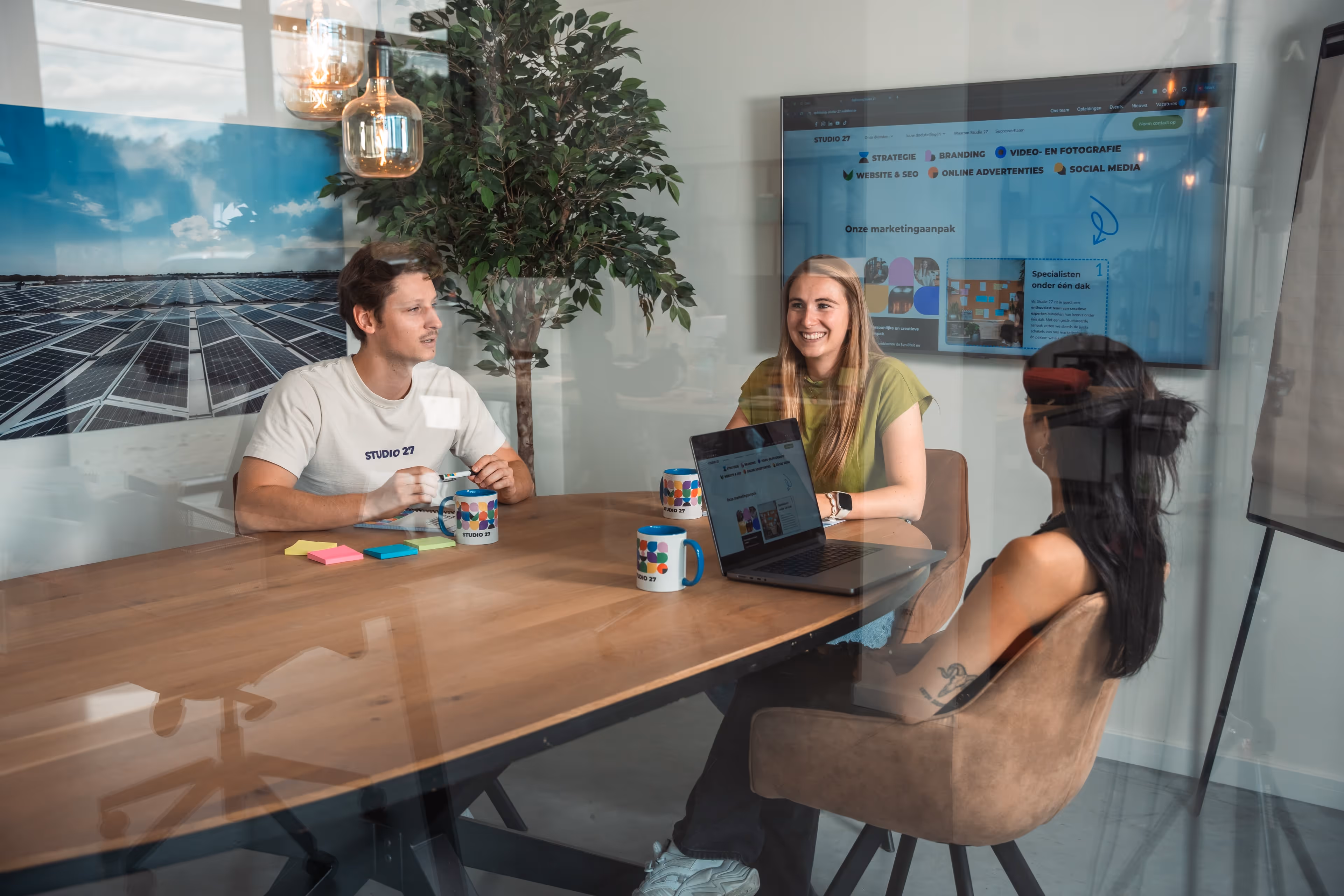 Drie mensen zitten rond een houten tafel in een kantoorruimte met laptop, mokken en notitieblokken, in gesprek met een scherm en een foto van zonnepanelen op de achtergrond.