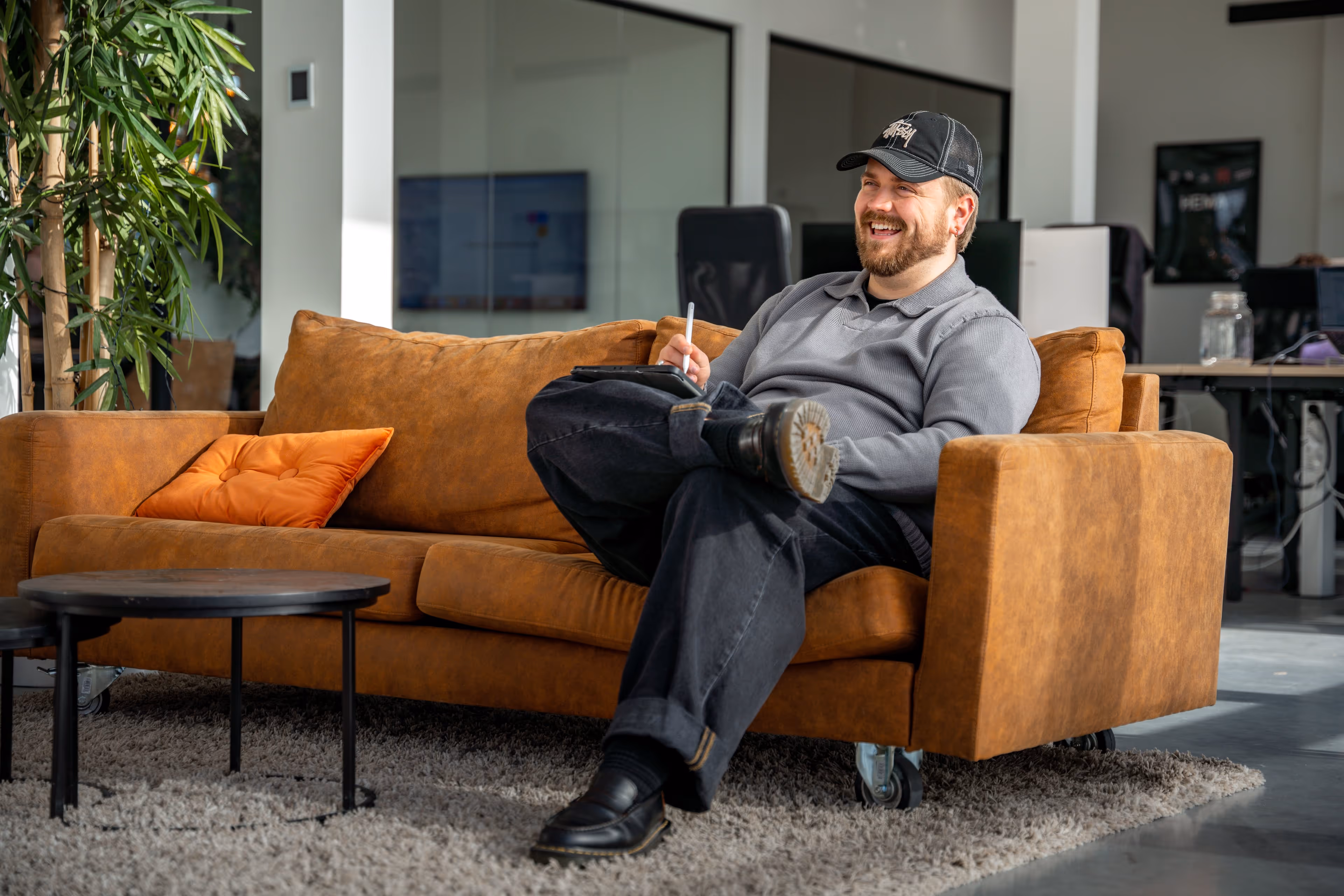 Man zittend op een bruine leren bank in een kantooromgeving, lachend en schrijvend op een tablet.