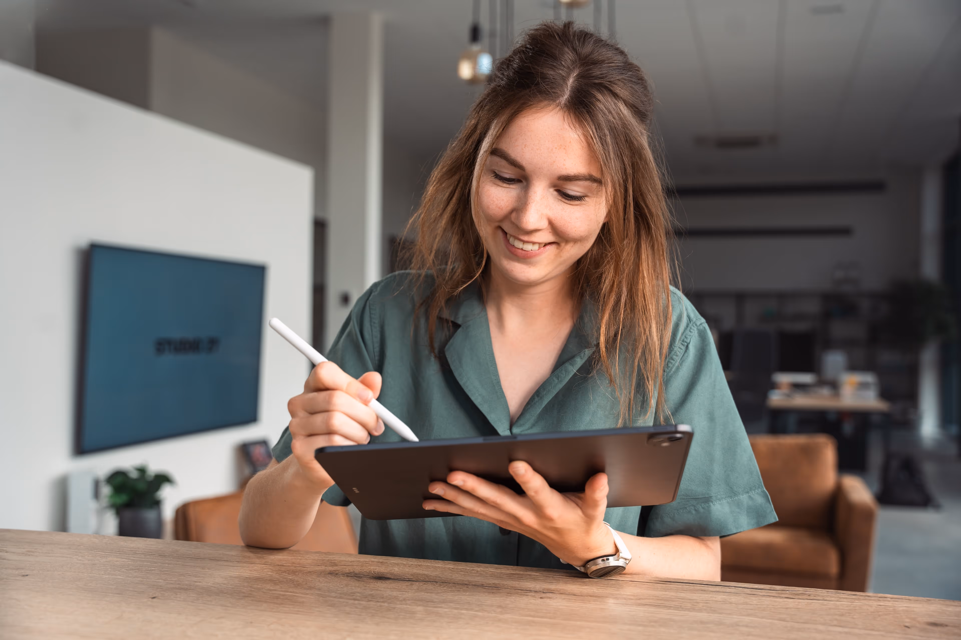 Vrouw glimlachend terwijl ze met een stylus op een tablet werkt aan een houten tafel in een moderne kantooromgeving.