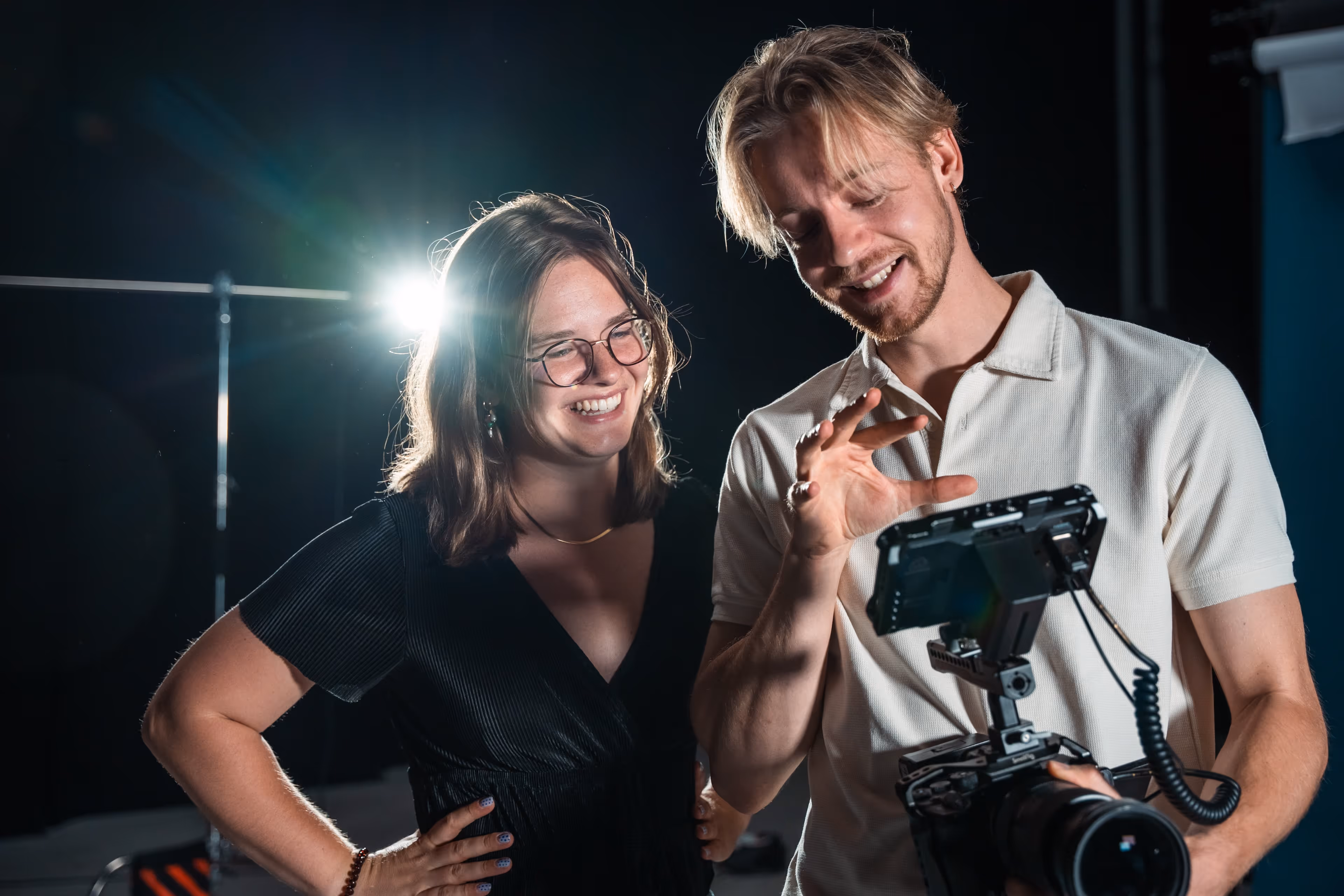 Twee lachende mensen bekijken samen beelden op een professionele filmcamera met monitor in een studio met donkere achtergrond.