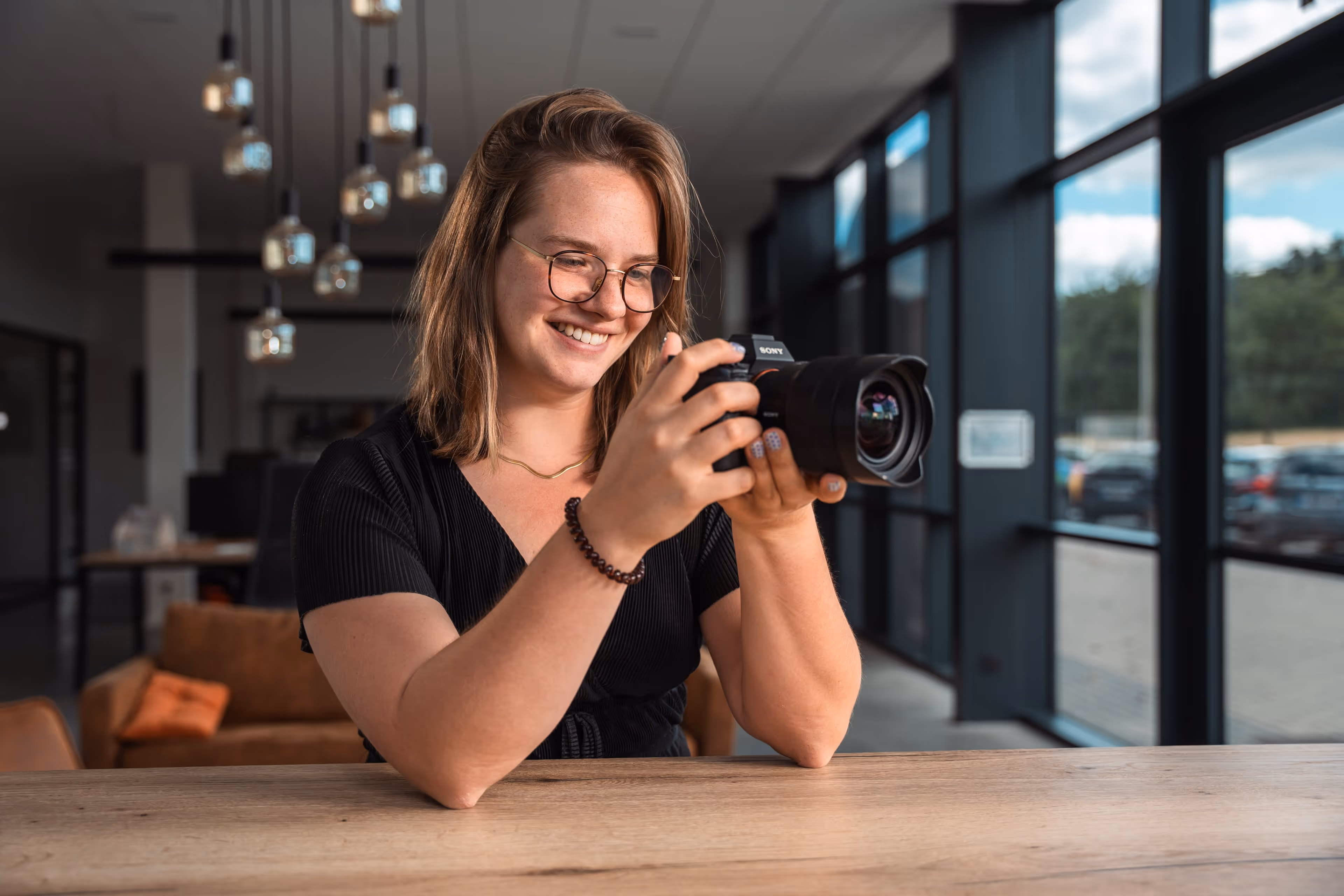 Vrouw met bril glimlacht terwijl ze een professionele camera vasthoudt in een moderne kantoorruimte.