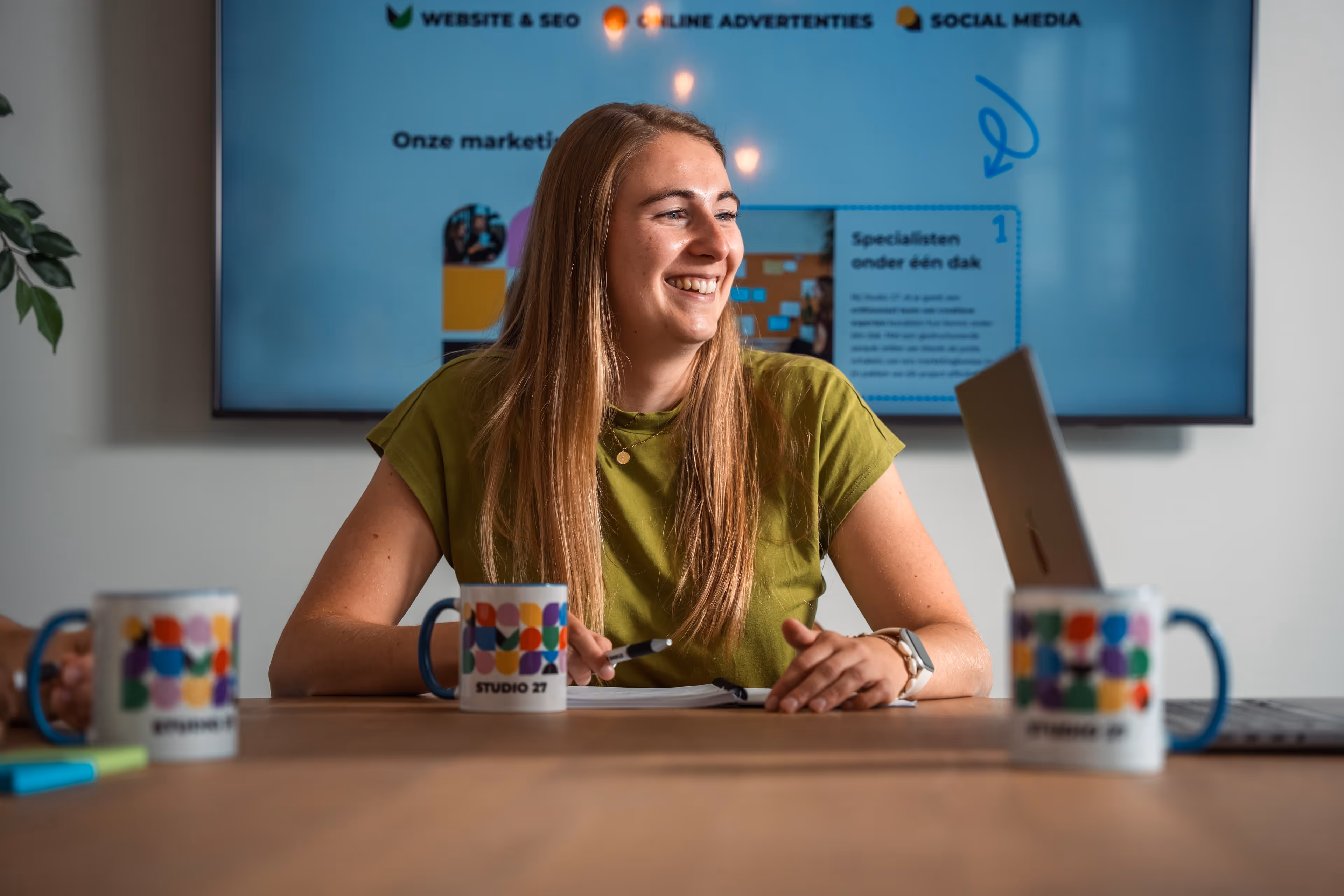 Vrouw met groene trui lacht zittend aan tafel met notitieboekje en pen, achtergrond met scherm en Studio 27 mokken op tafel.
