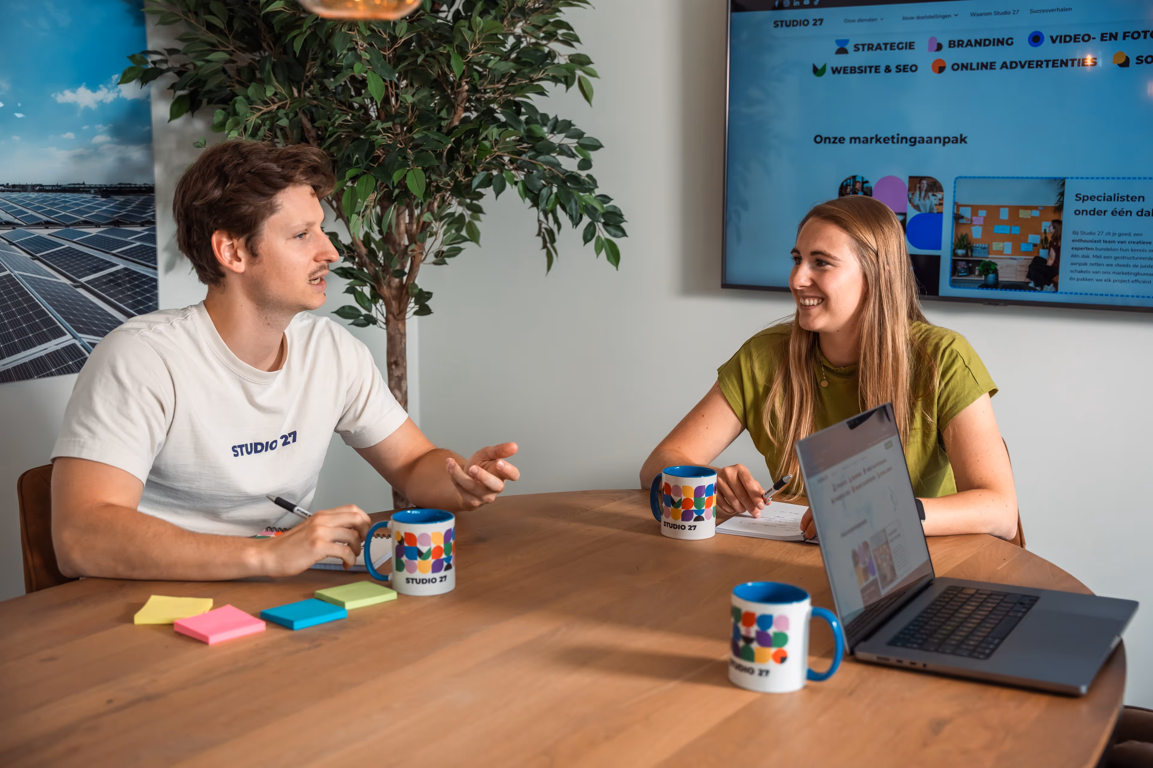 Twee mensen in een kantoorruimte zitten aan een tafel met Studio 27 mokken, notities en een laptop, terwijl ze een gesprek voeren.