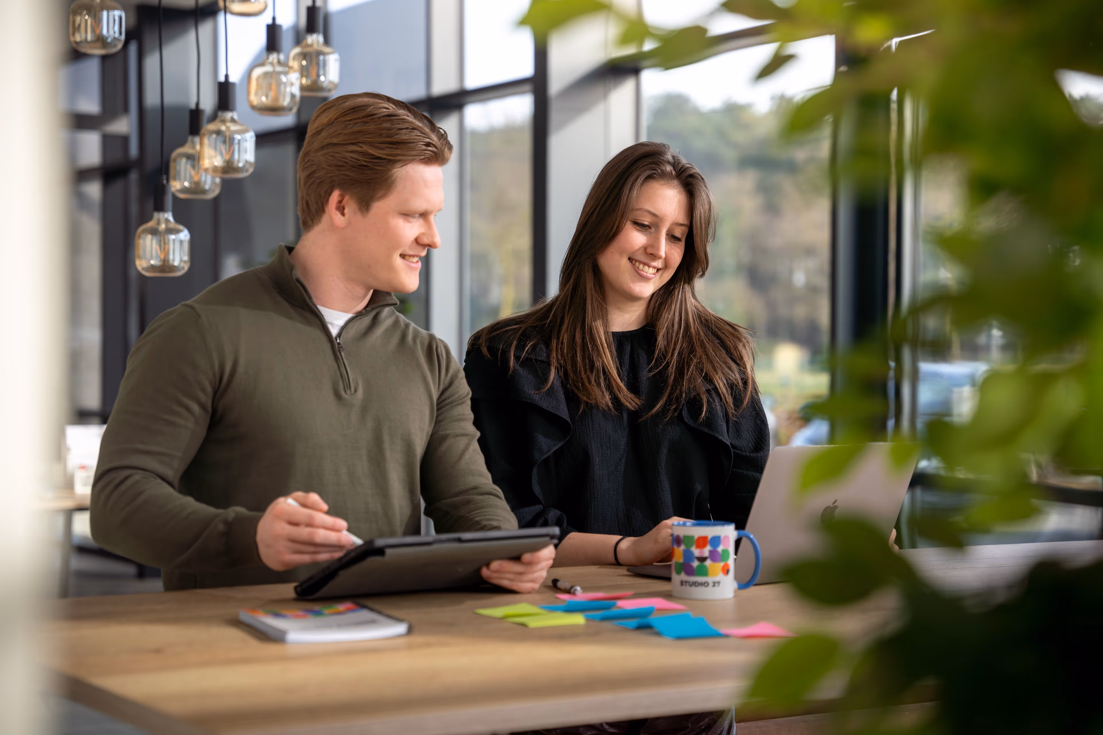 Twee jonge volwassenen werken samen aan een tafel met een laptop, tablet en kleurrijke plaknotities in een lichte, moderne kantoorruimte.