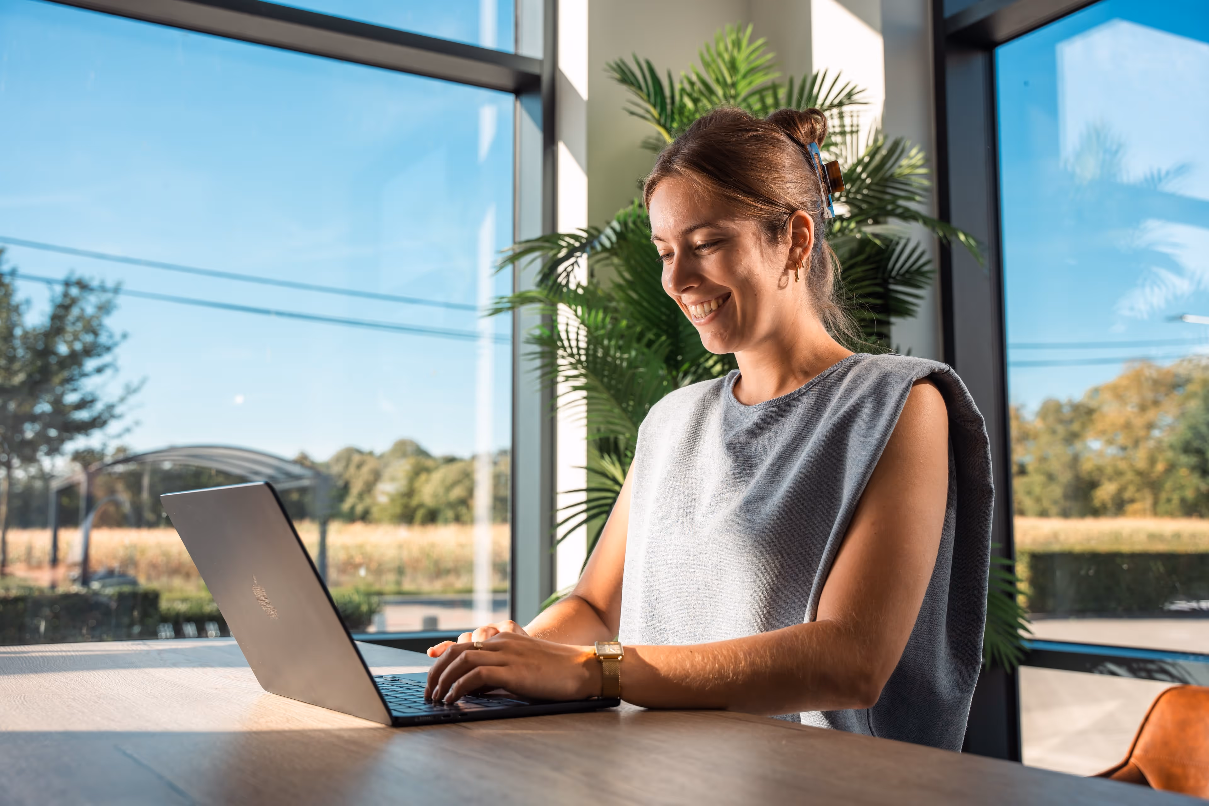 Vrouw die glimlachend op een laptop werkt aan een tafel in een ruimte met grote ramen en groene planten.