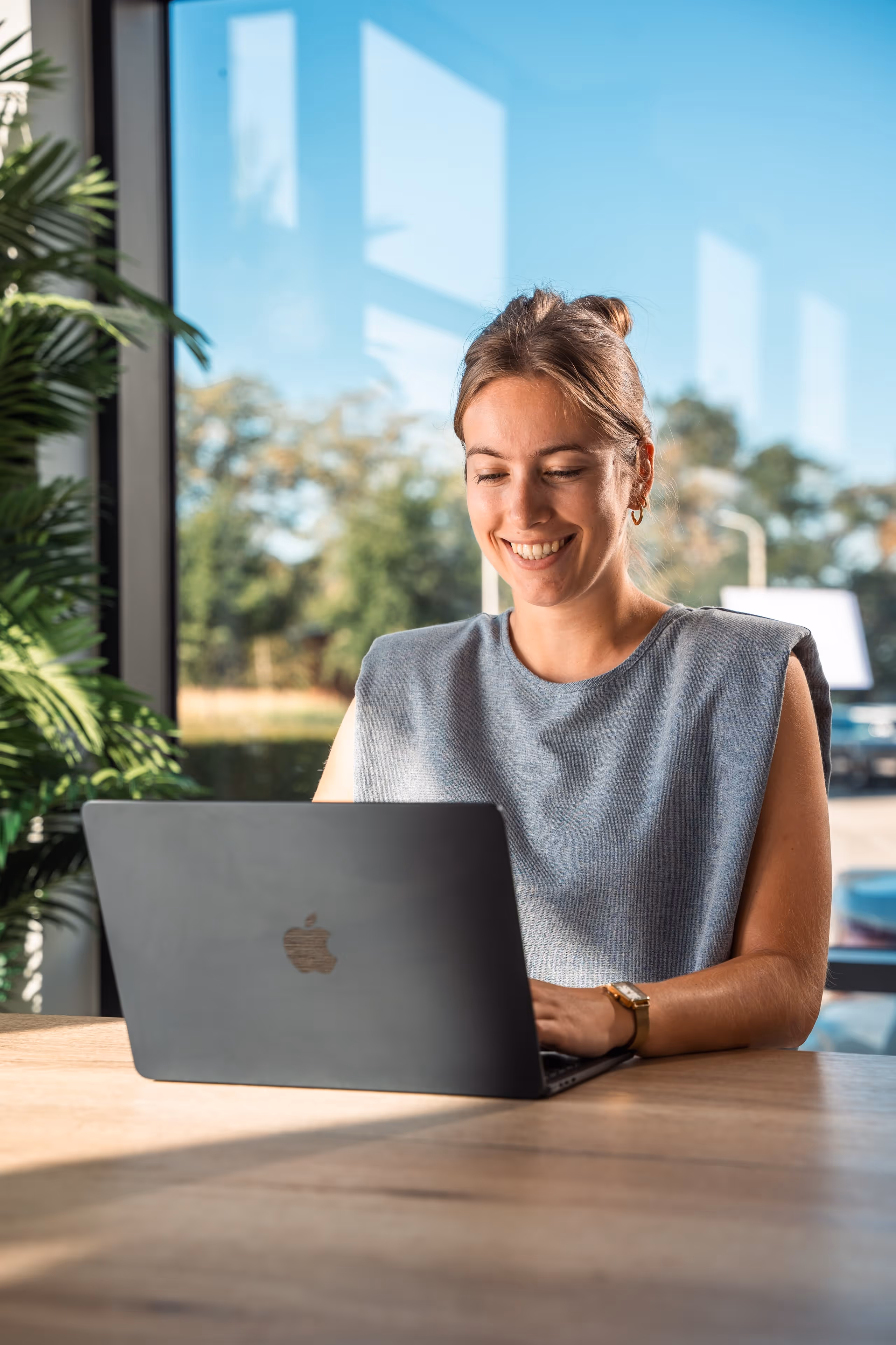 Vrouw die glimlacht terwijl ze aan een bureau werkt op een laptop, met een groot raam en planten op de achtergrond.