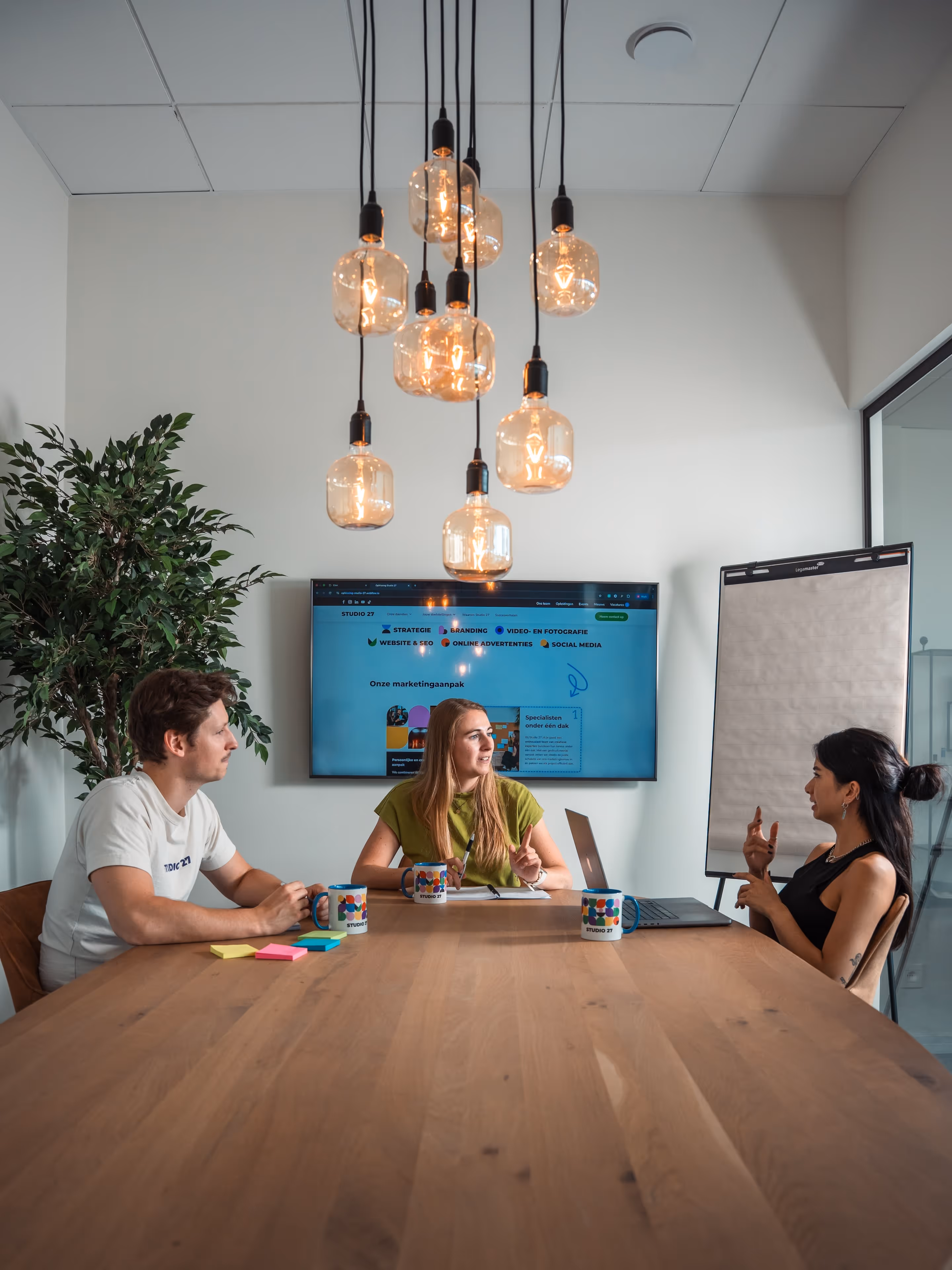 Drie mensen in een moderne vergaderruimte zitten aan een houten tafel met koffiekopjes, een laptop en een tv-scherm met marketinginhoud aan de muur.
