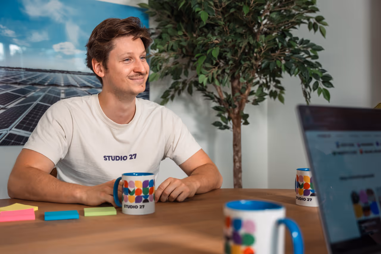 Jonge man in wit Studio 27 T-shirt zit aan tafel met kleurrijke mokken, laptop en post-its, met een plant en een afbeelding van zonnepanelen op de achtergrond.