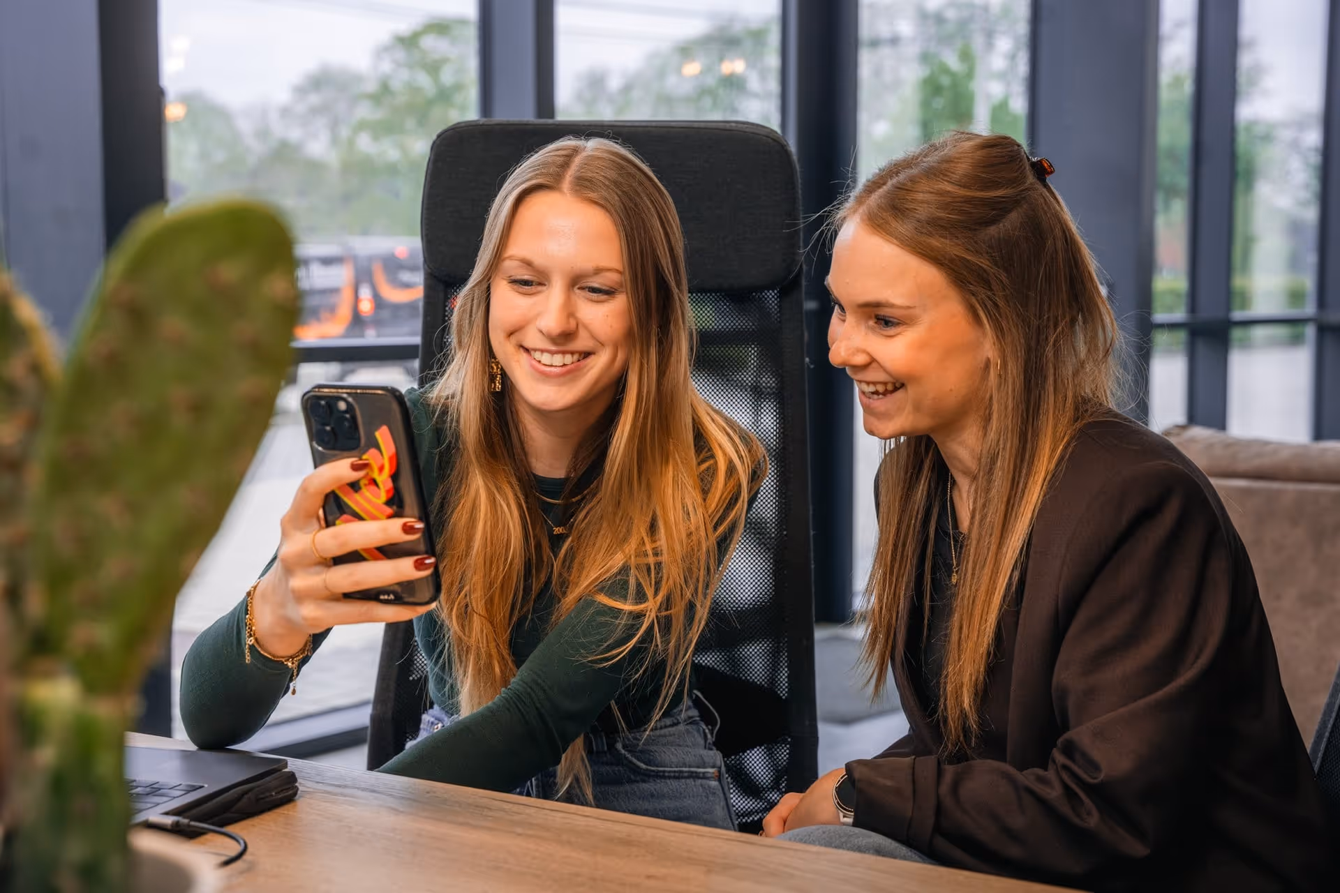 Twee vrouwen zitten samen aan een tafel, glimlachend terwijl ze naar een telefoon kijken.