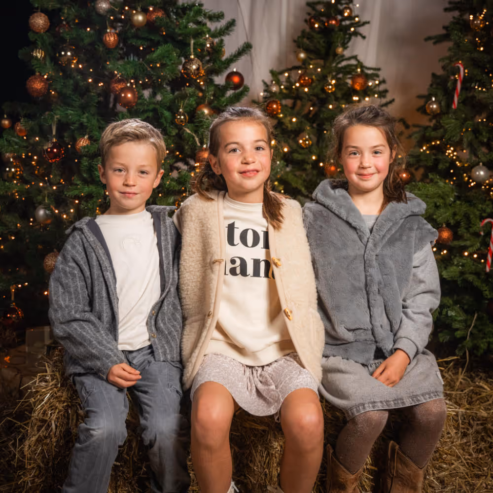 Drie kinderen zitten samen op een hooibaal voor versierde kerstbomen met lichtjes en ornamenten.