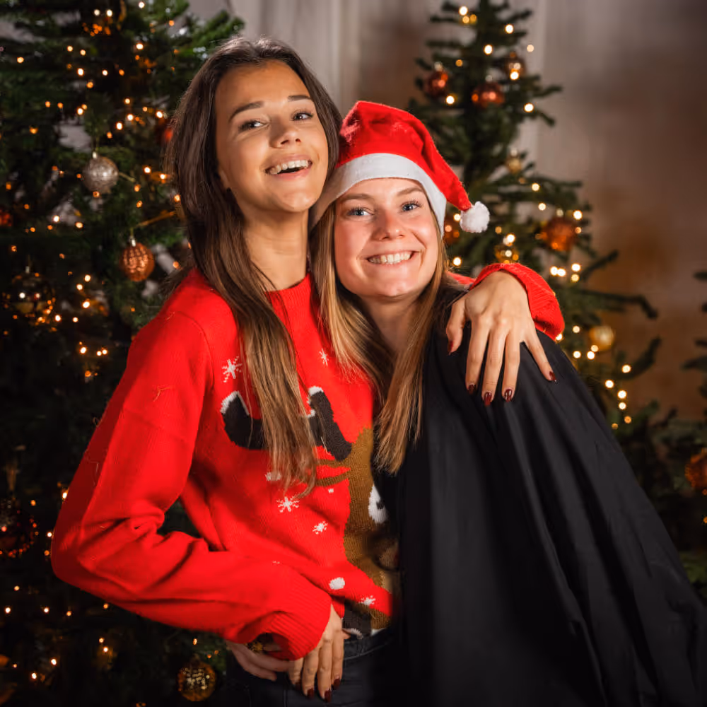 Twee vrouwen omhelzen elkaar lachend voor versierde kerstbomen, de ene draagt een rode trui met rendier, de ander een kerstmanmuts.