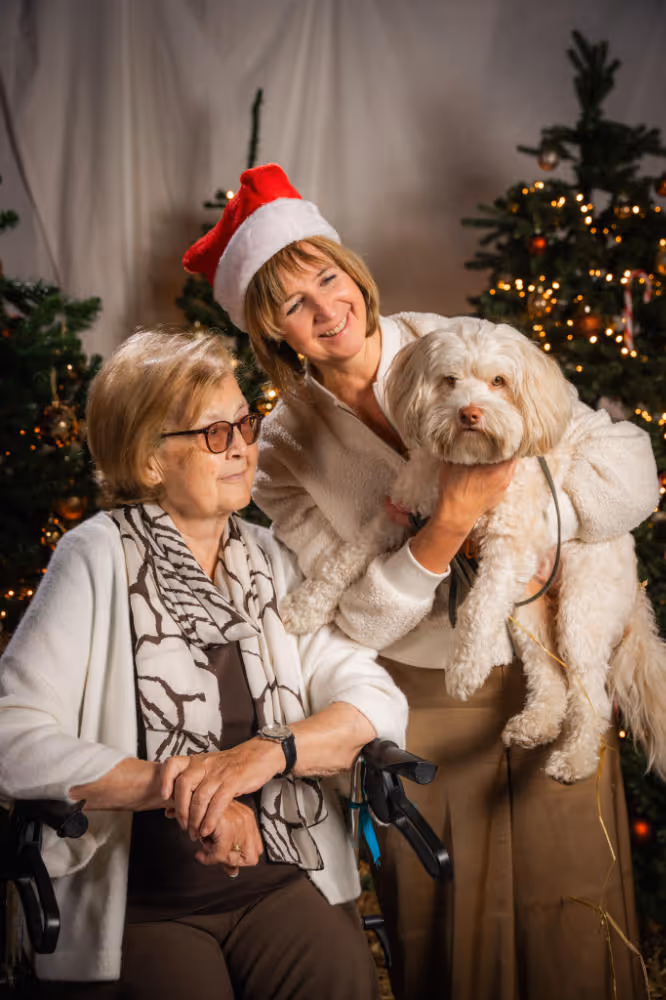 Een oudere vrouw in een rolstoel naast een jongere vrouw met een kerstmanmuts die een kleine witte hond vasthoudt, met versierde kerstbomen op de achtergrond.