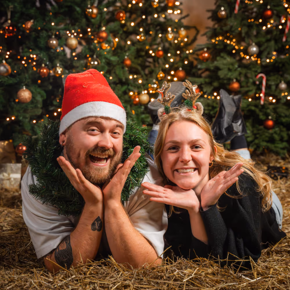 Man met een kerstmuts en vrouw met rendieroren liggen samen op stro voor verlichte kerstbomen.