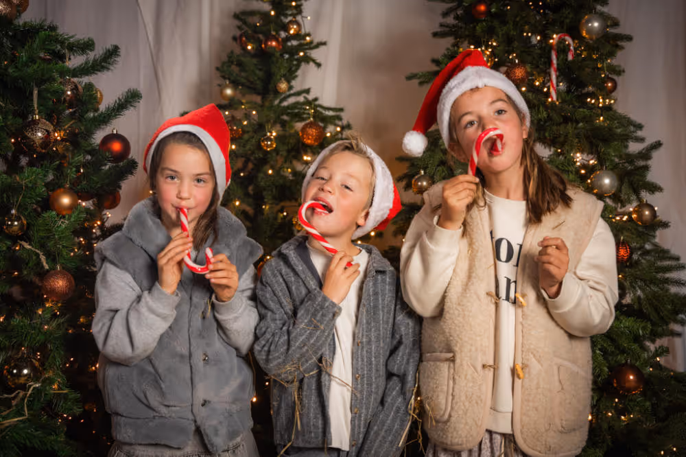 Drie kinderen met kerstmutsen die zuurstokken vasthouden voor versierde kerstbomen.