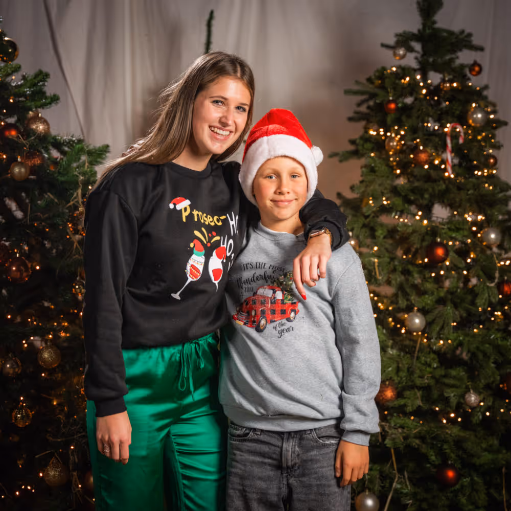 Vrouw en jongen in feestelijke truien poseren samen voor verlichte kerstbomen.