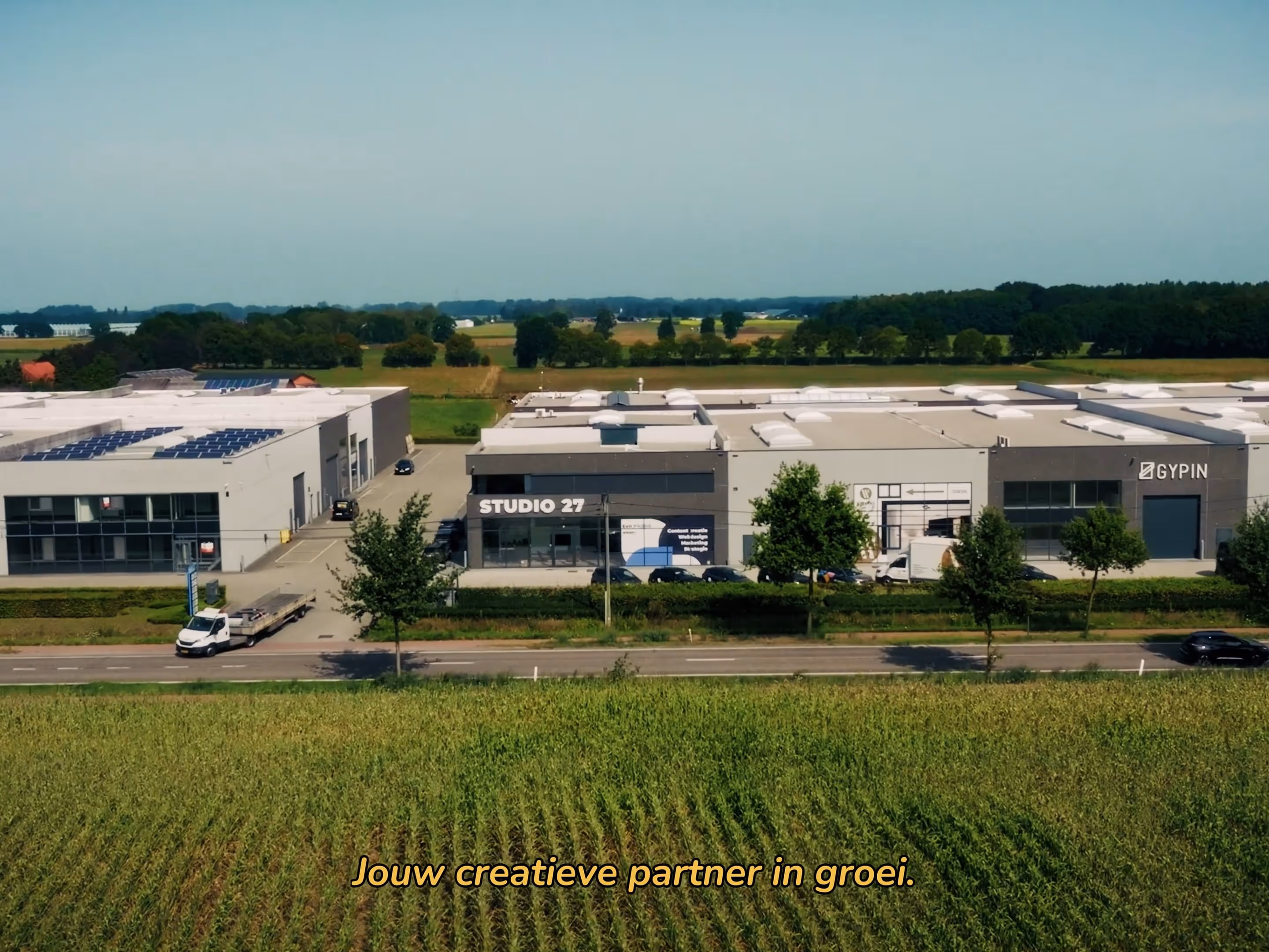 Luchtfoto van bedrijventerrein met moderne gebouwen, waaronder één met de naam Studio 27, omgeven door groen en landbouwvelden.