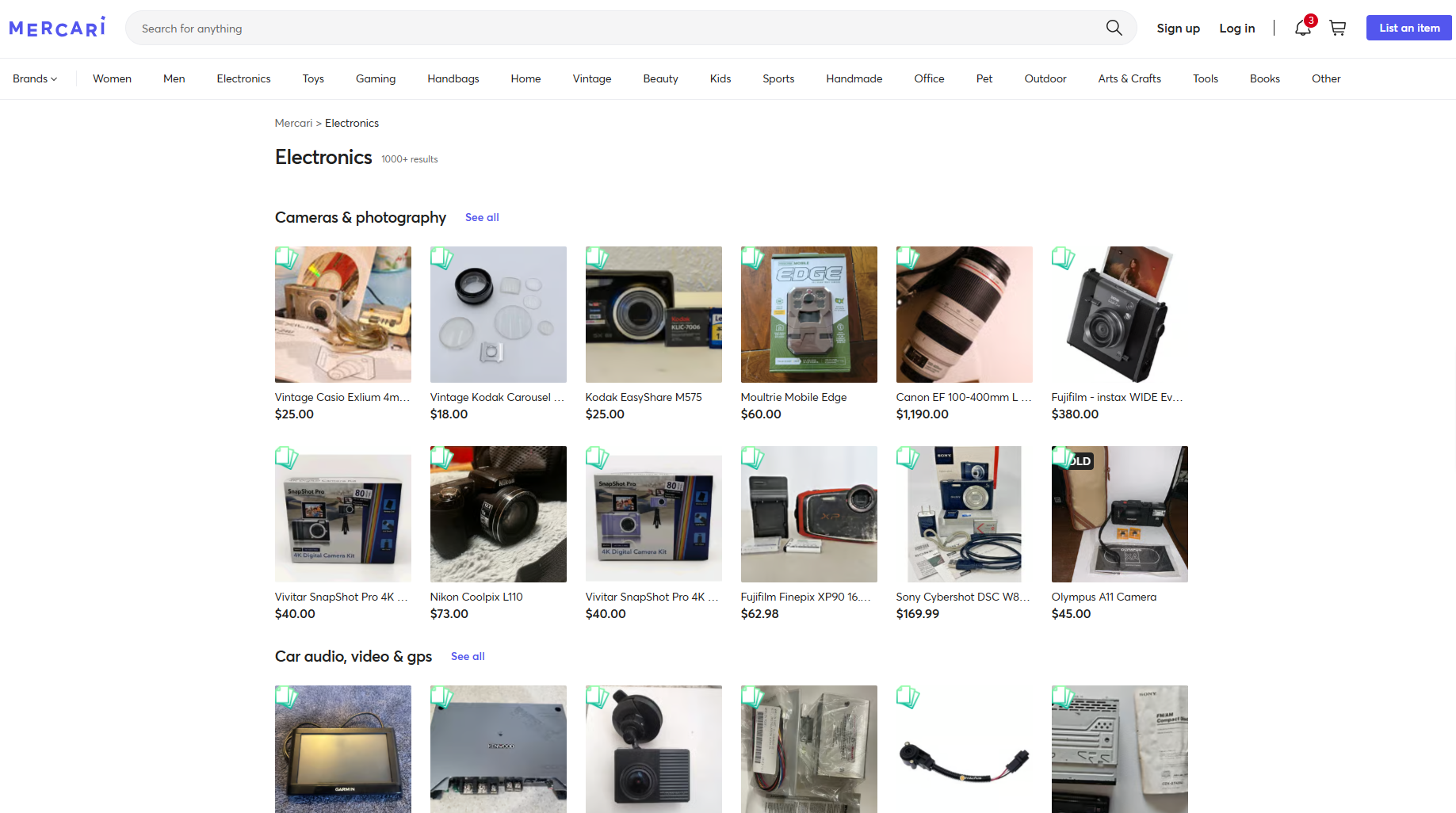 Here's the alt-text: Screenshot of Mercari's Electronics category page (1,000+ results) showing two sections: "Cameras & photography" and "Car audio, video & GPS." The cameras grid displays 12 listings, including a Canon EF 100–400mm L lens ($1,190), Fujifilm Instax WIDE ($380), Sony Cybershot DSC W8 ($169.99), and several budget point-and-shoot cameras ranging from $18–$73. The Mercari navigation bar is visible at the top.