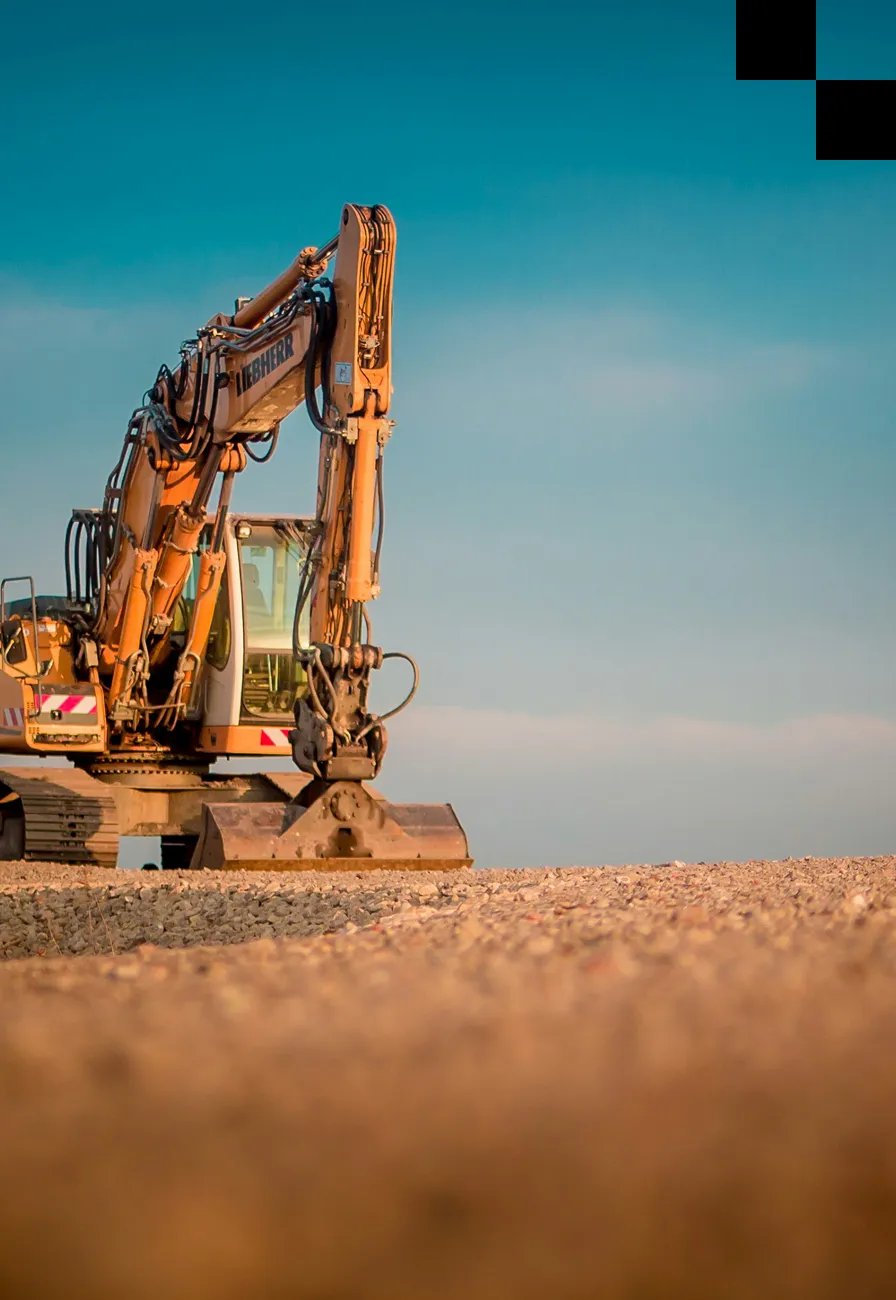 A large yellow excavator sits on a gravel surface under a clear blue sky, with its arm and bucket lowered, ready for construction work.