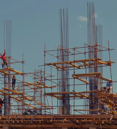 Several construction workers stand on scaffolding, working with steel rebar frameworks and wooden planks on a building site under a clear blue sky.