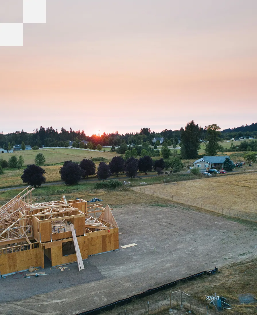 A partially constructed wooden house frame stands on a dirt lot in a rural area at sunset, with trees, open fields, and distant houses visible under a colorful sky.