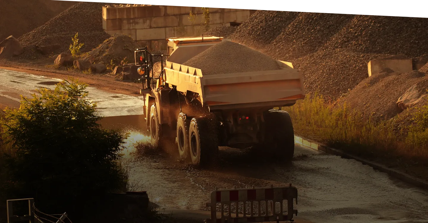 A large dump truck loaded with gravel drives on a muddy road at a construction or mining site, surrounded by dirt piles and greenery in warm sunlight.