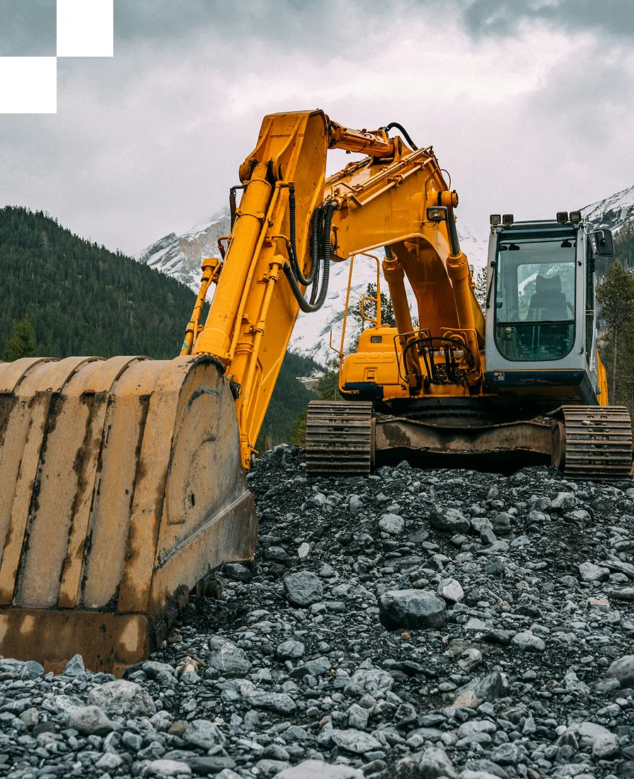 A large yellow excavator sits on rocky terrain with a mountainous landscape and cloudy sky in the background.