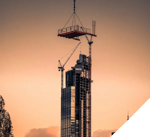 A construction crane lifts a large platform to the top of a tall, modern skyscraper at sunset. The building has glass windows and the word eavis displayed near the top.