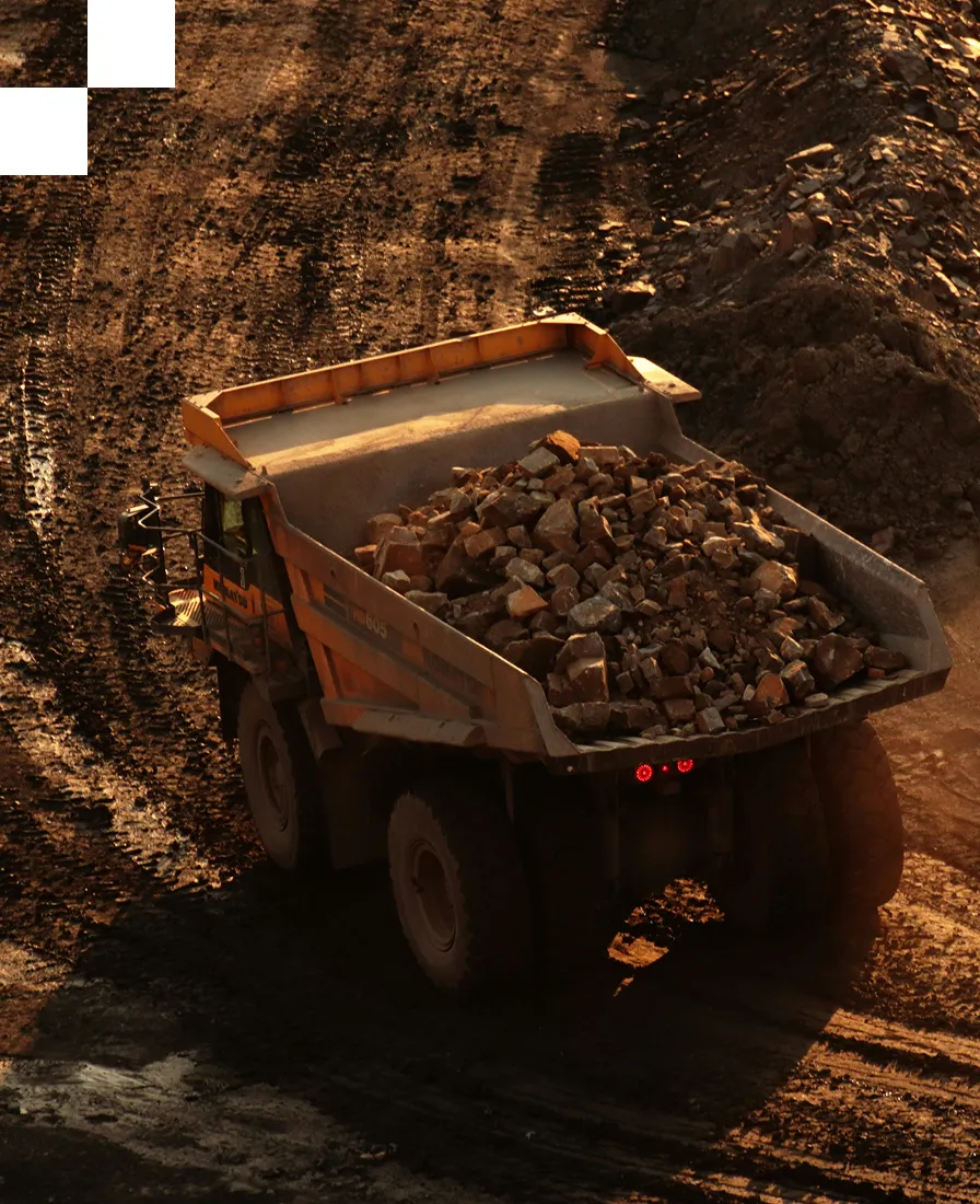 A large yellow dump truck loaded with rocks drives along a dirt road at a mining or construction site during sunset, casting long shadows on the ground.