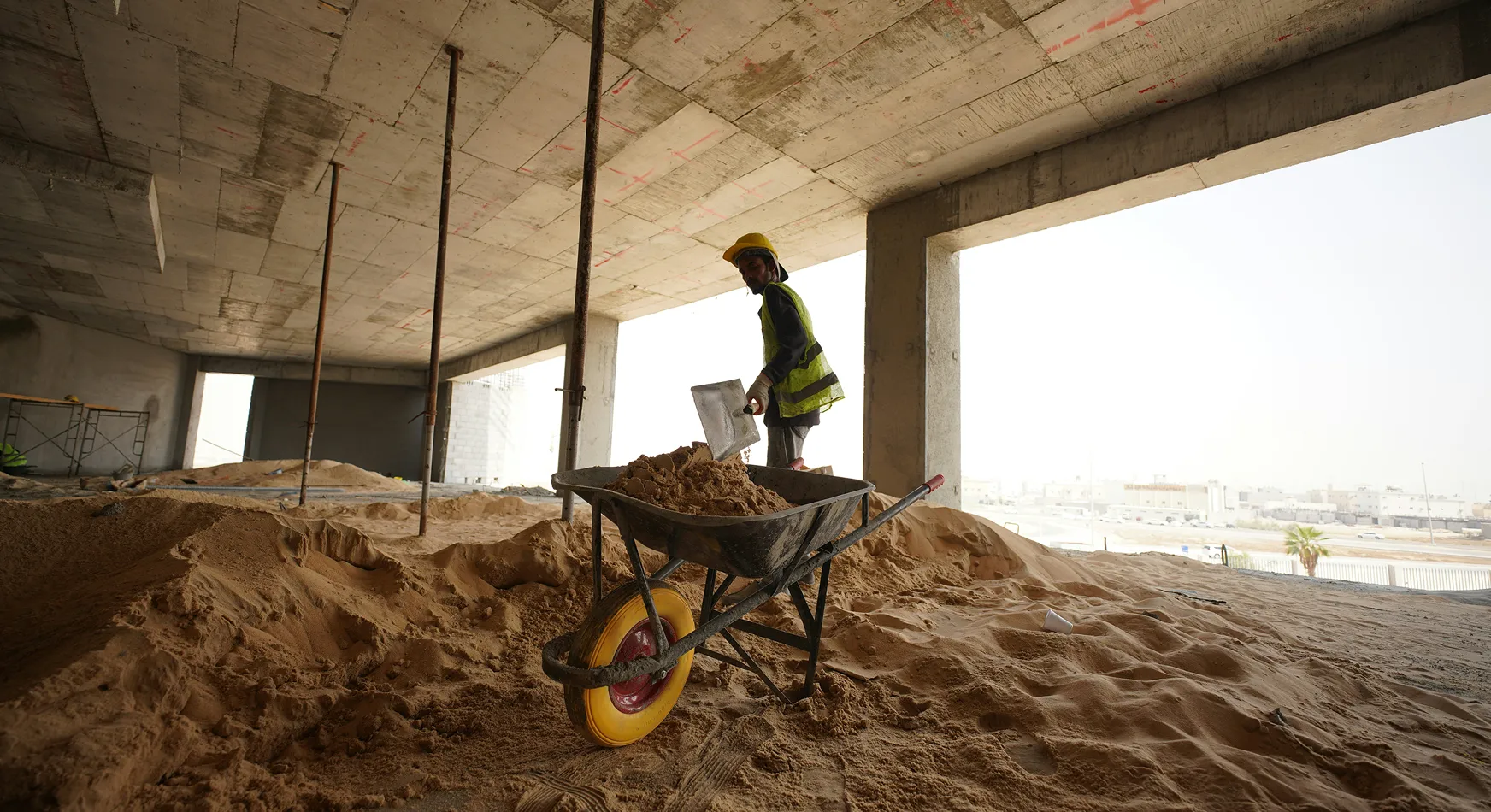 A construction worker in a yellow hard hat and vest stands near a wheelbarrow filled with sand in an unfinished building, with large windows and a view of a city outside.
