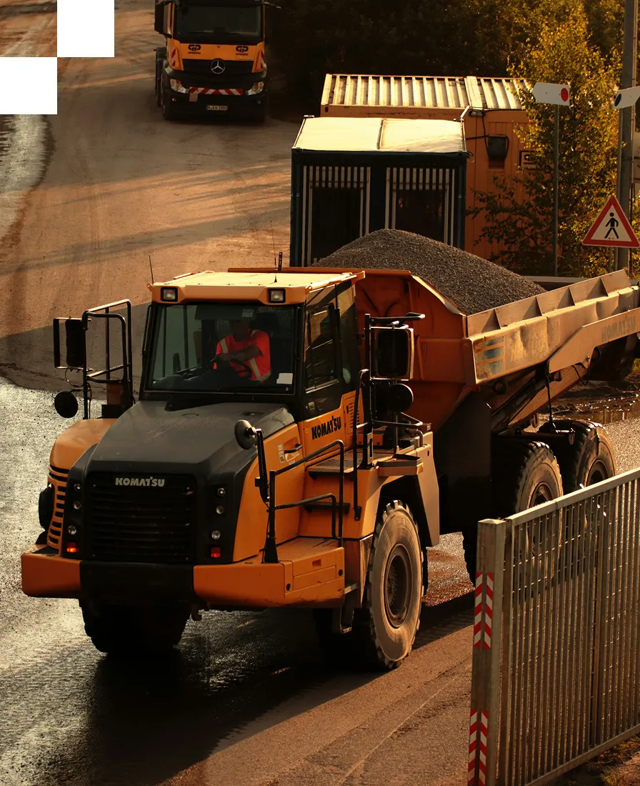 A large yellow Komatsu dump truck loaded with gravel drives through an industrial site near a security gate and a guard booth, with another truck and trees visible in the background.