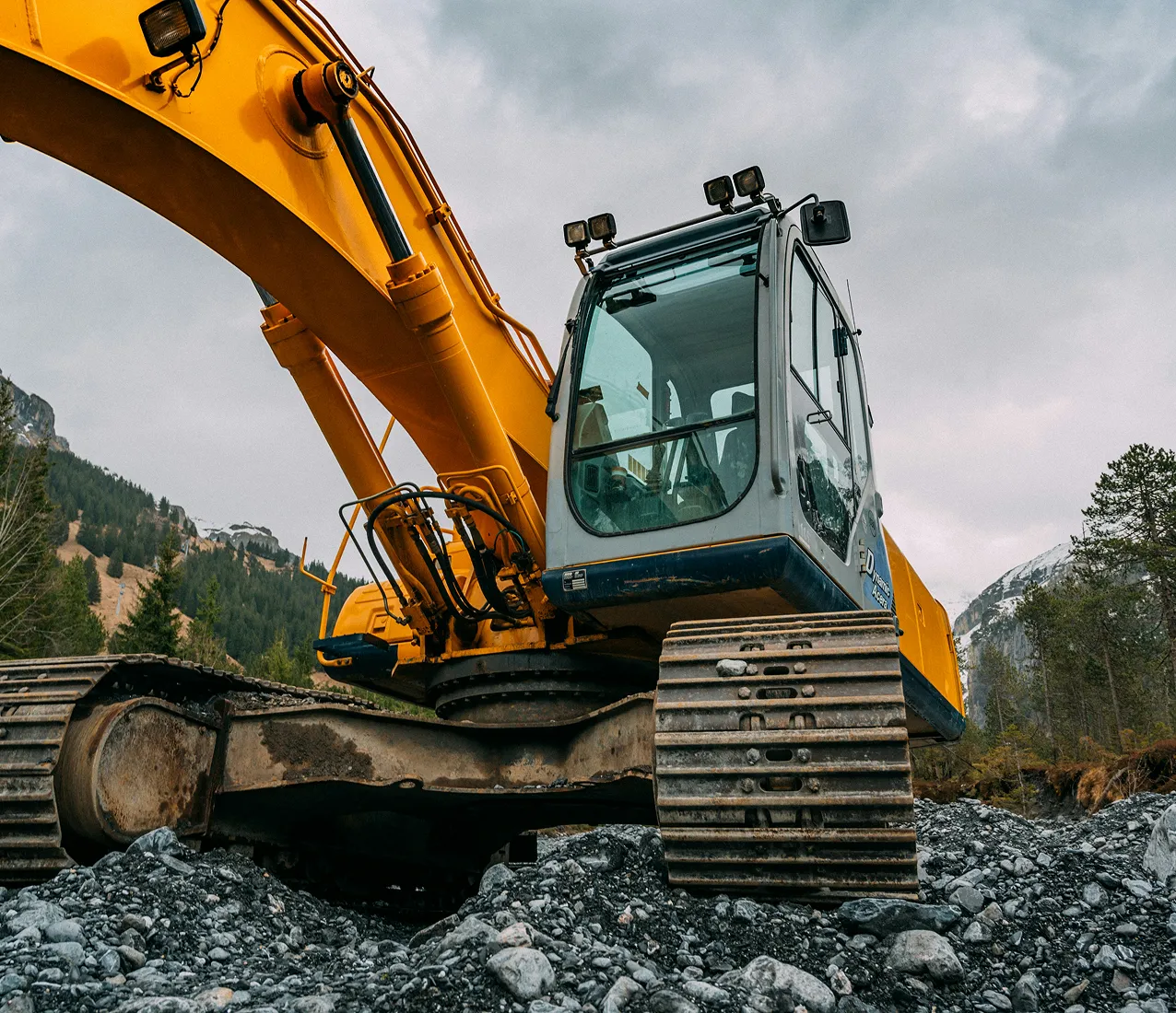 A yellow excavator with large metal tracks sits on rocky ground outdoors, surrounded by trees and mountains under a cloudy sky.
