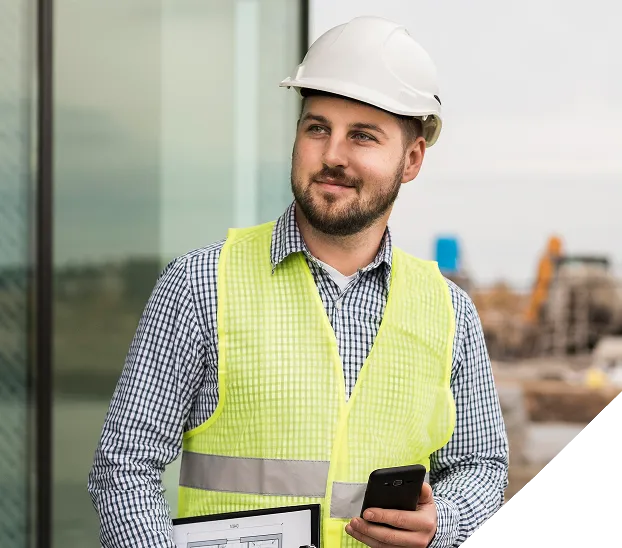 A construction worker wearing a white helmet and neon safety vest stands with arms crossed, smiling in front of a large excavator at a construction site.