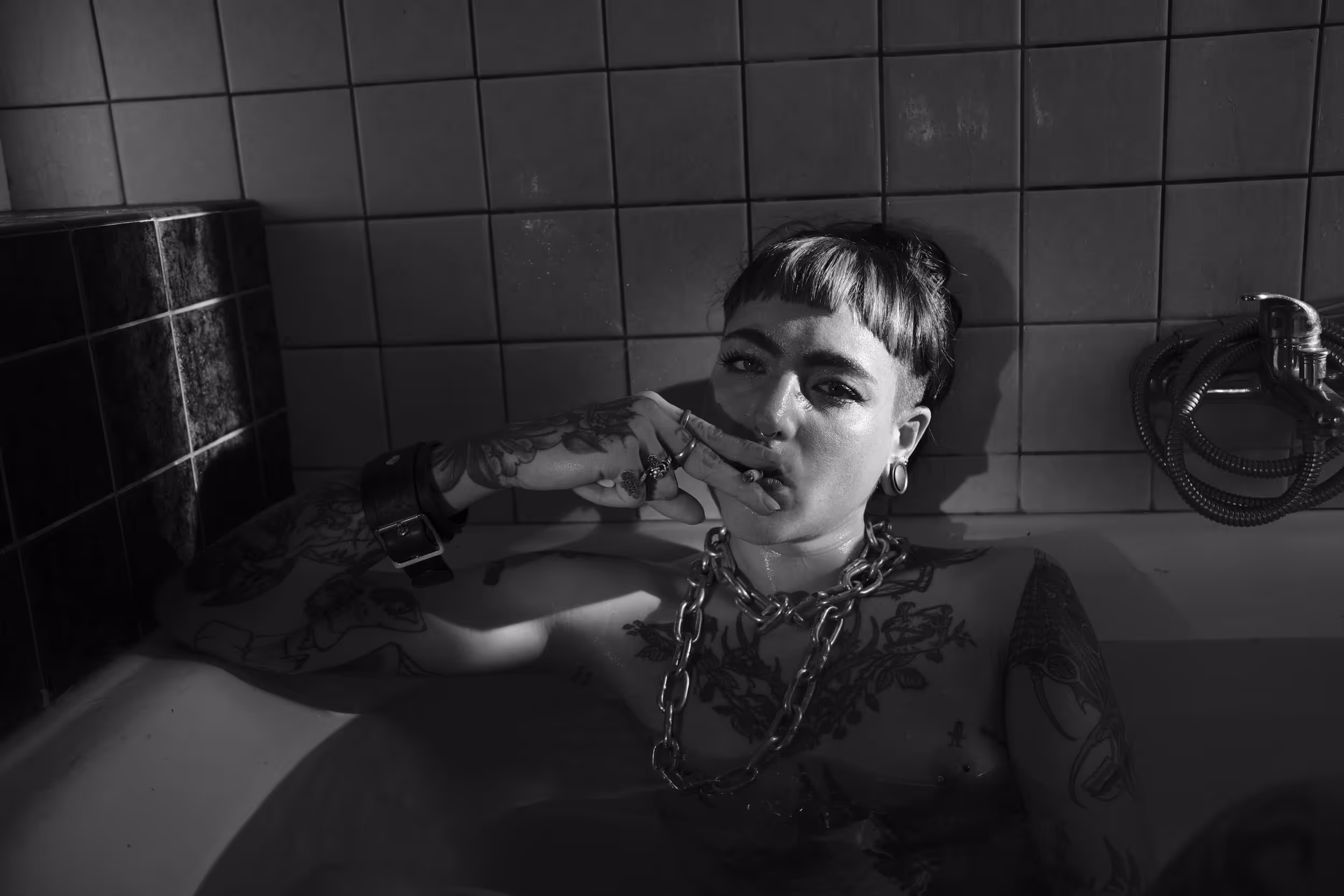 Femme tatouée dans une baignoire, regard fixe et cigarette à la main, dans une lumière contrastée.