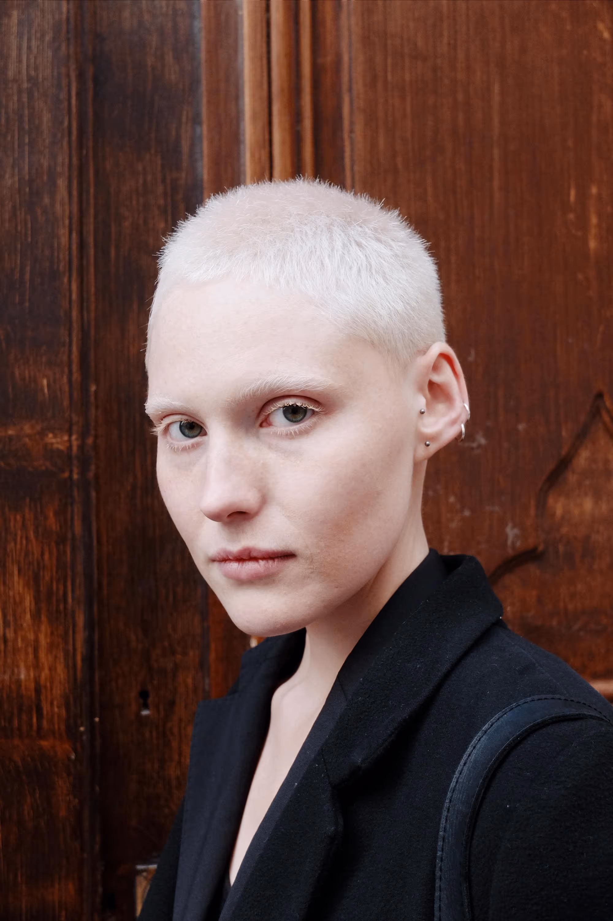 Jeune femme aux cheveux très courts et blonds platine, regard direct vers l’objectif, devant une porte en bois.