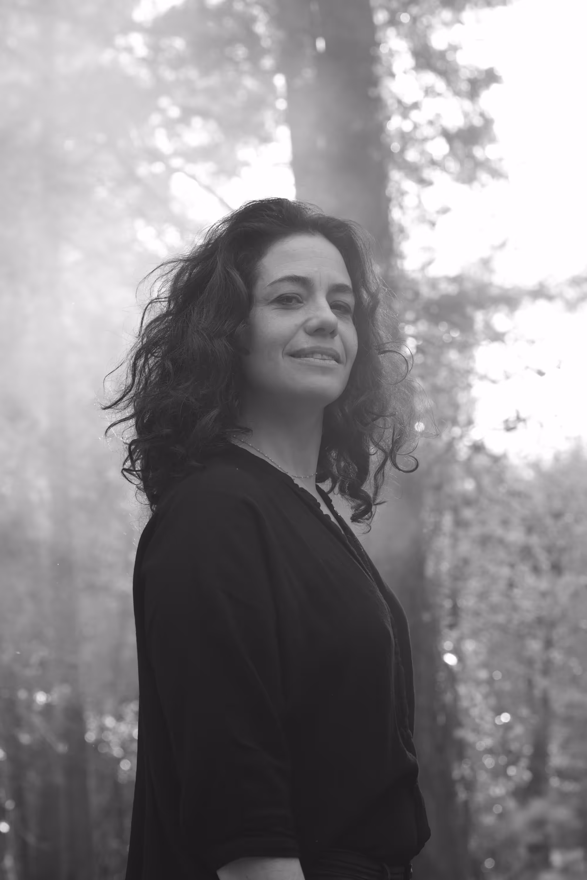 Femme aux cheveux bouclés debout en pleine forêt, lumière douce en arrière-plan.
