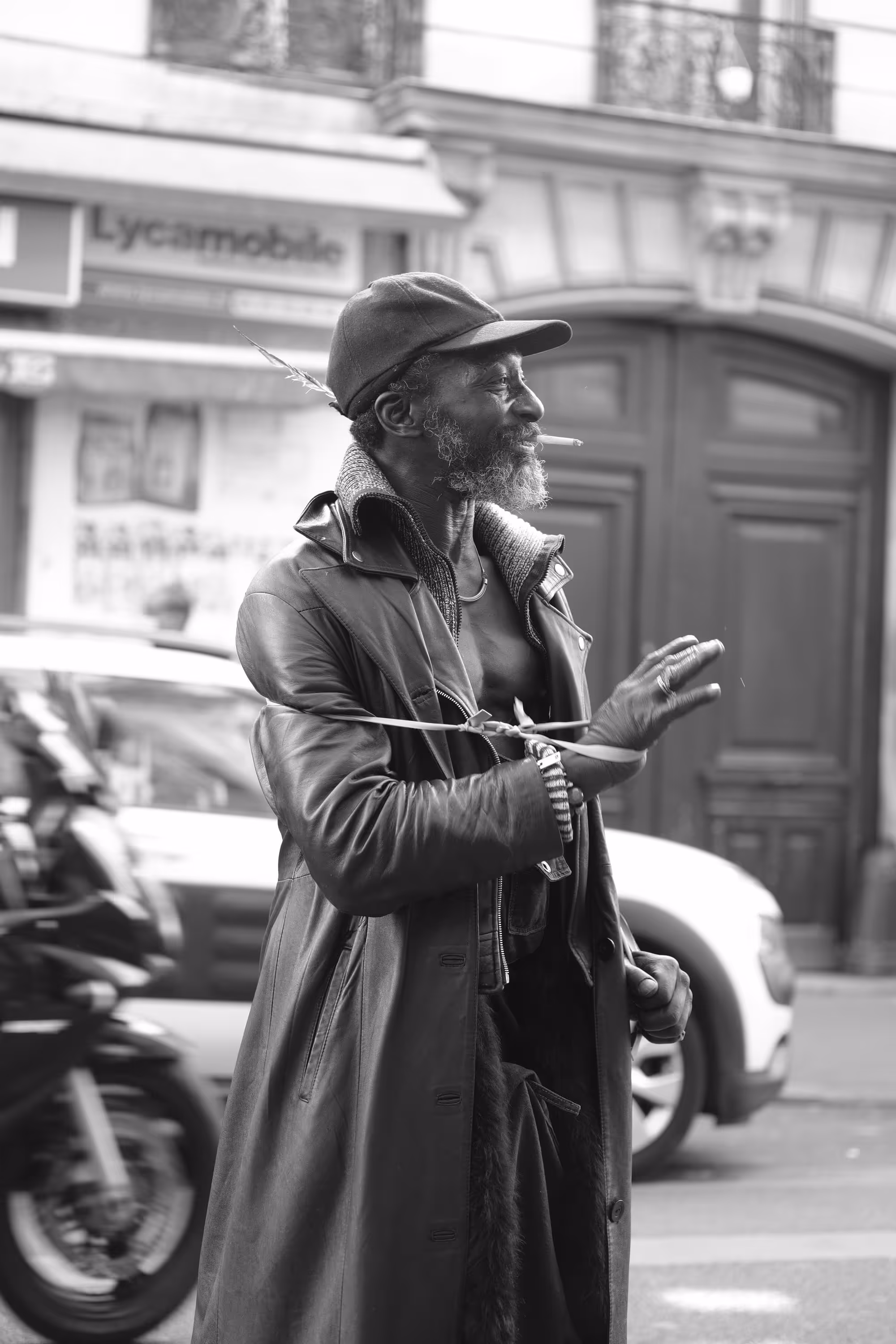 Homme vêtu d’un long manteau en cuir, cigarette à la bouche, dans une rue animée.