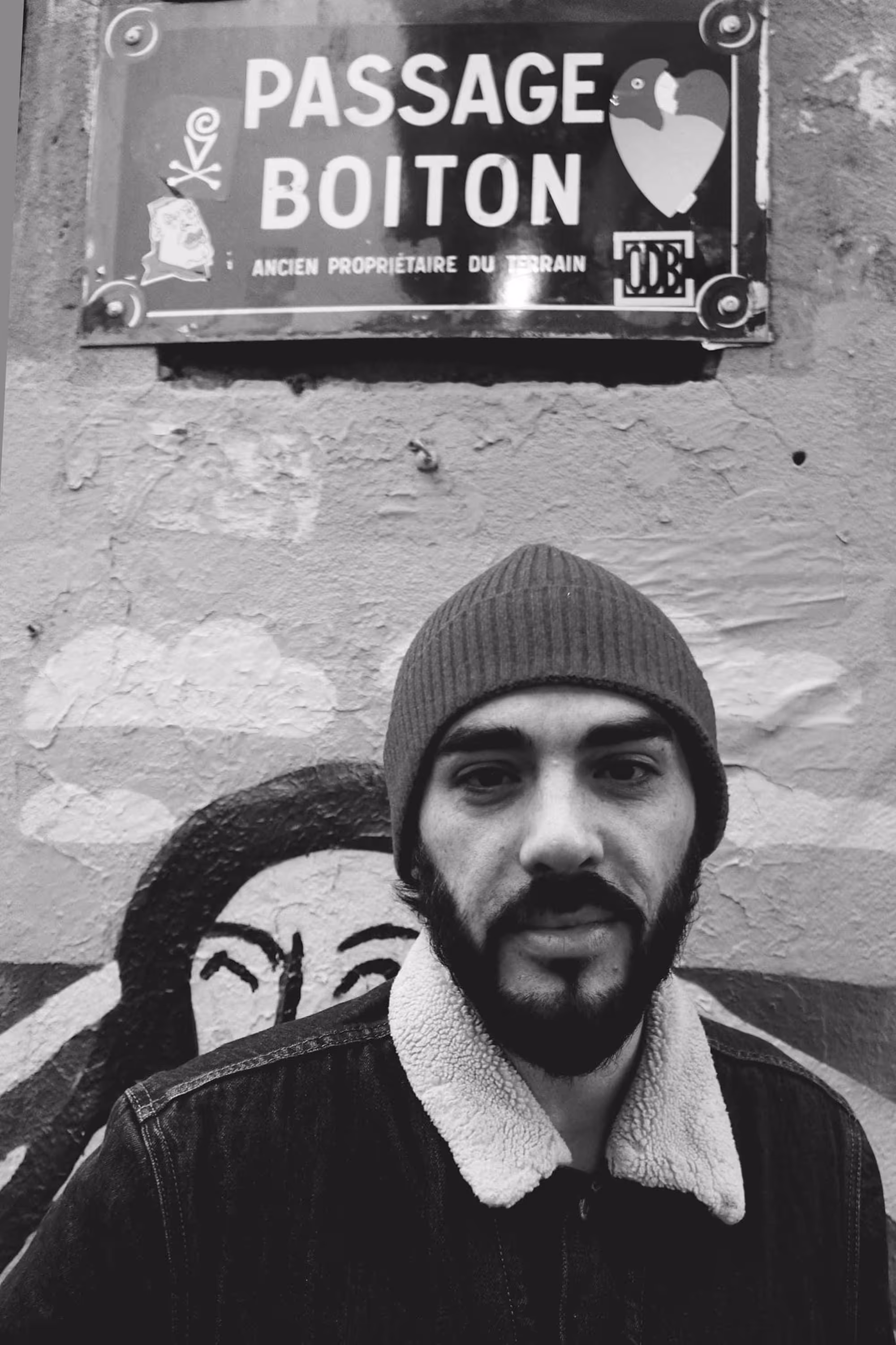 Homme barbu en bonnet, posé devant une fresque murale indiquant 'Passage Boiton'.