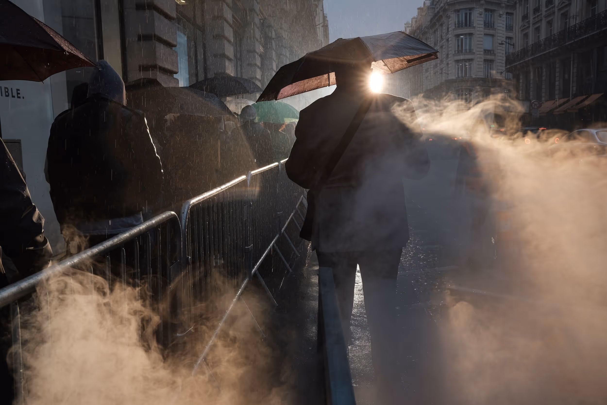 Homme sous un parapluie au coucher du soleil, entouré de vapeur dans une file d’attente sous la pluie.