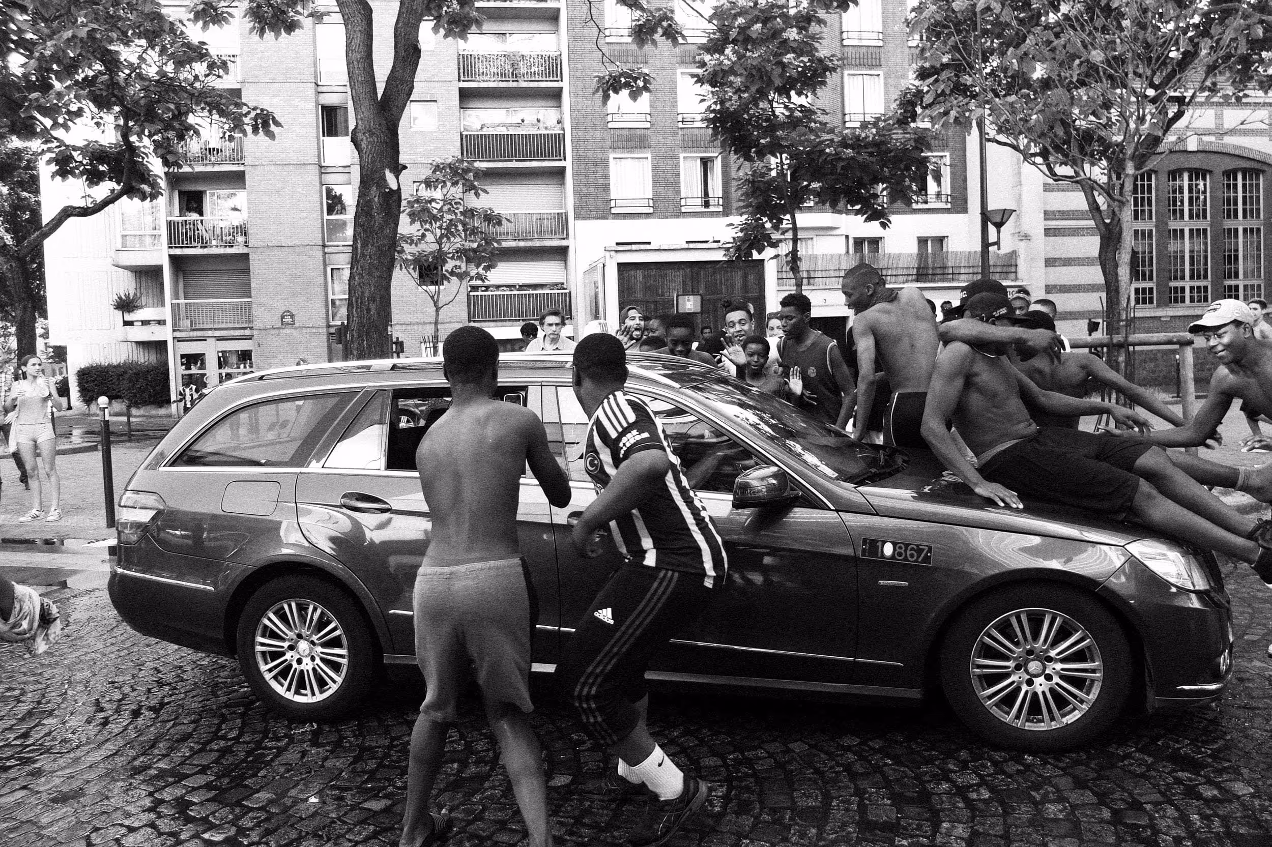 Adolescents autour et sur une voiture dans une rue pavée d’un quartier urbain en été.
