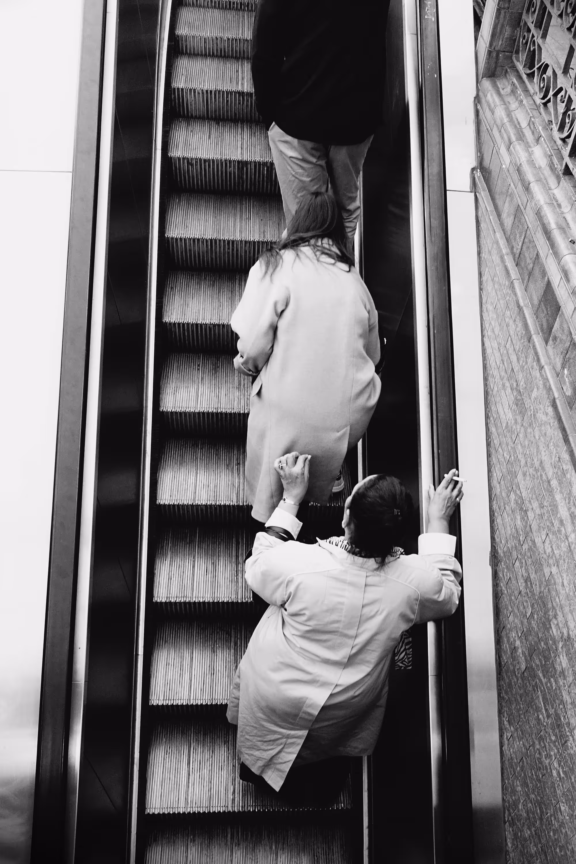 Vue en plongée d’une femme en train de gravir un escalator, une cigarette à la main.