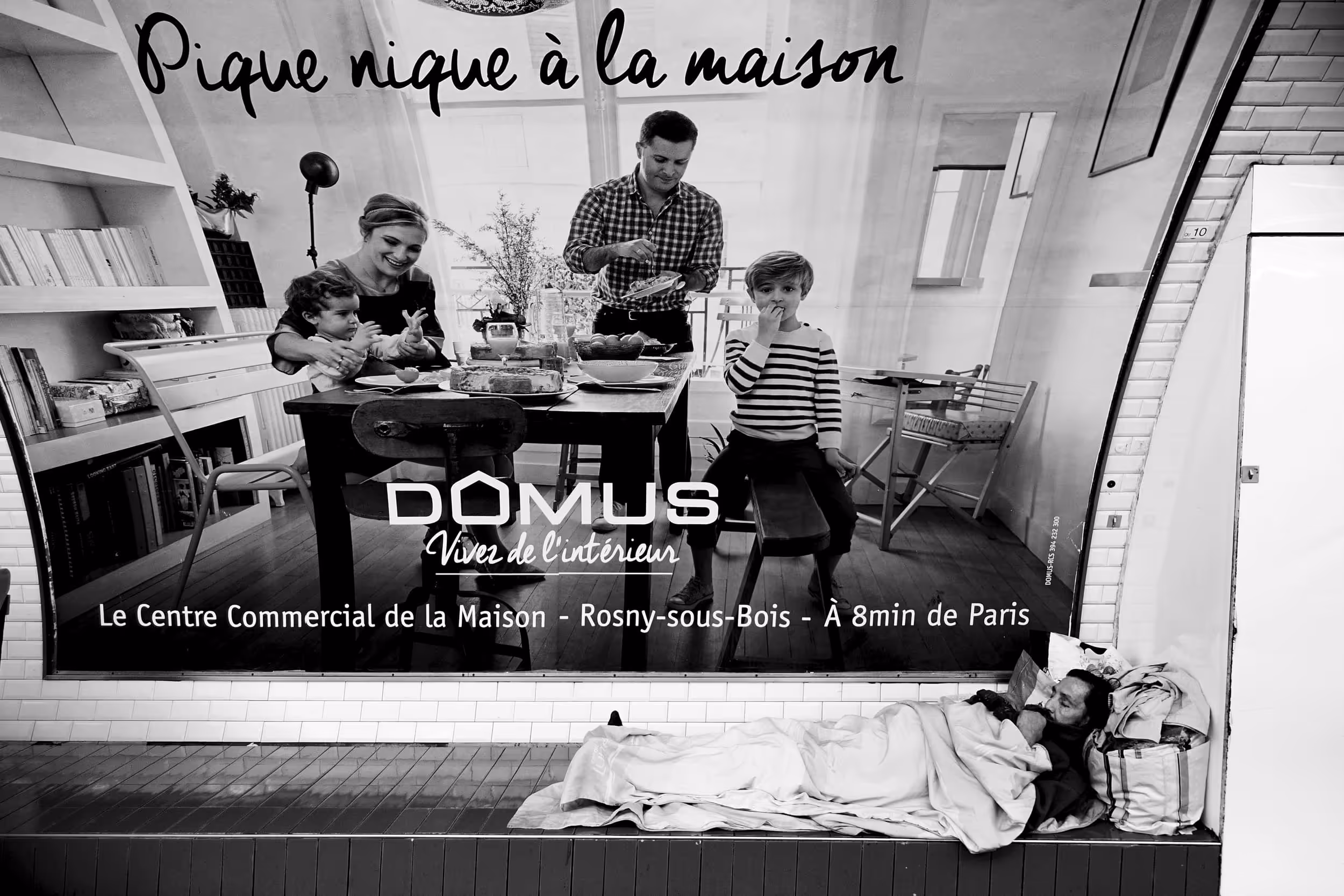 Un homme sans-abri dort enroulé dans une couverture sous une affiche publicitaire d’une famille attablée.