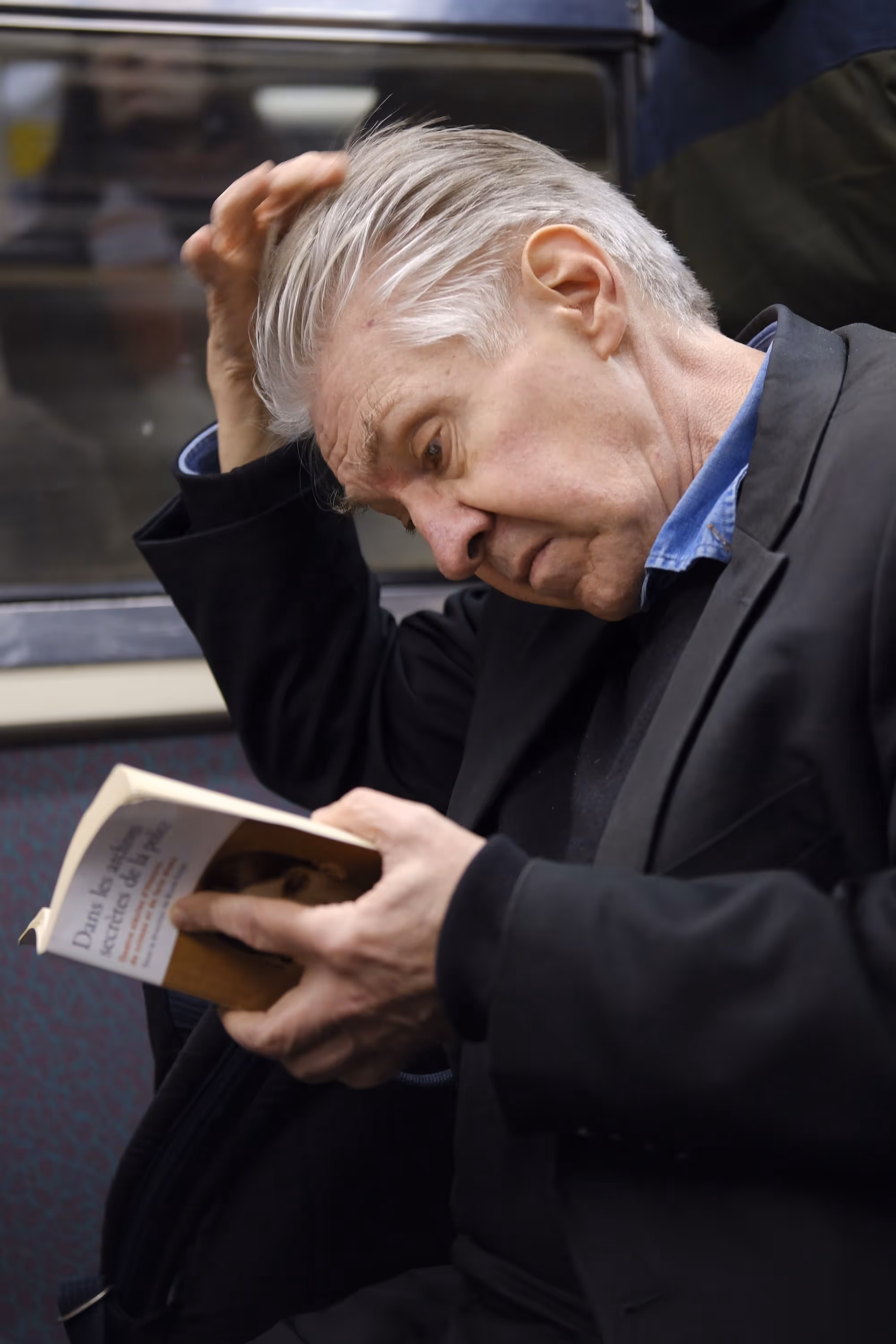 Homme âgé lisant un livre tout en se grattant la tête dans une rame de métro.