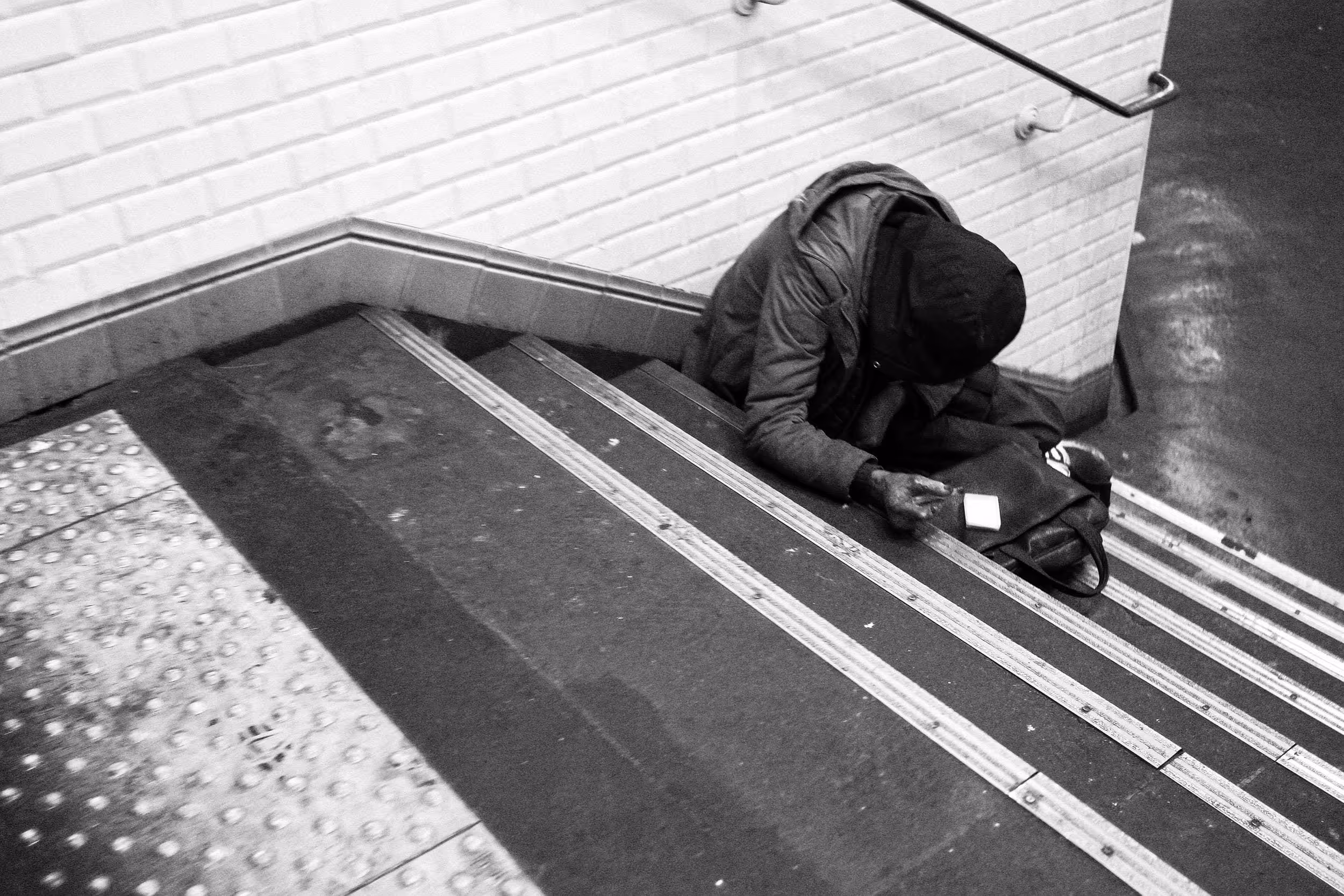 Une personne en situation de précarité est assise sur les marches d’un escalier de métro, main tendue.