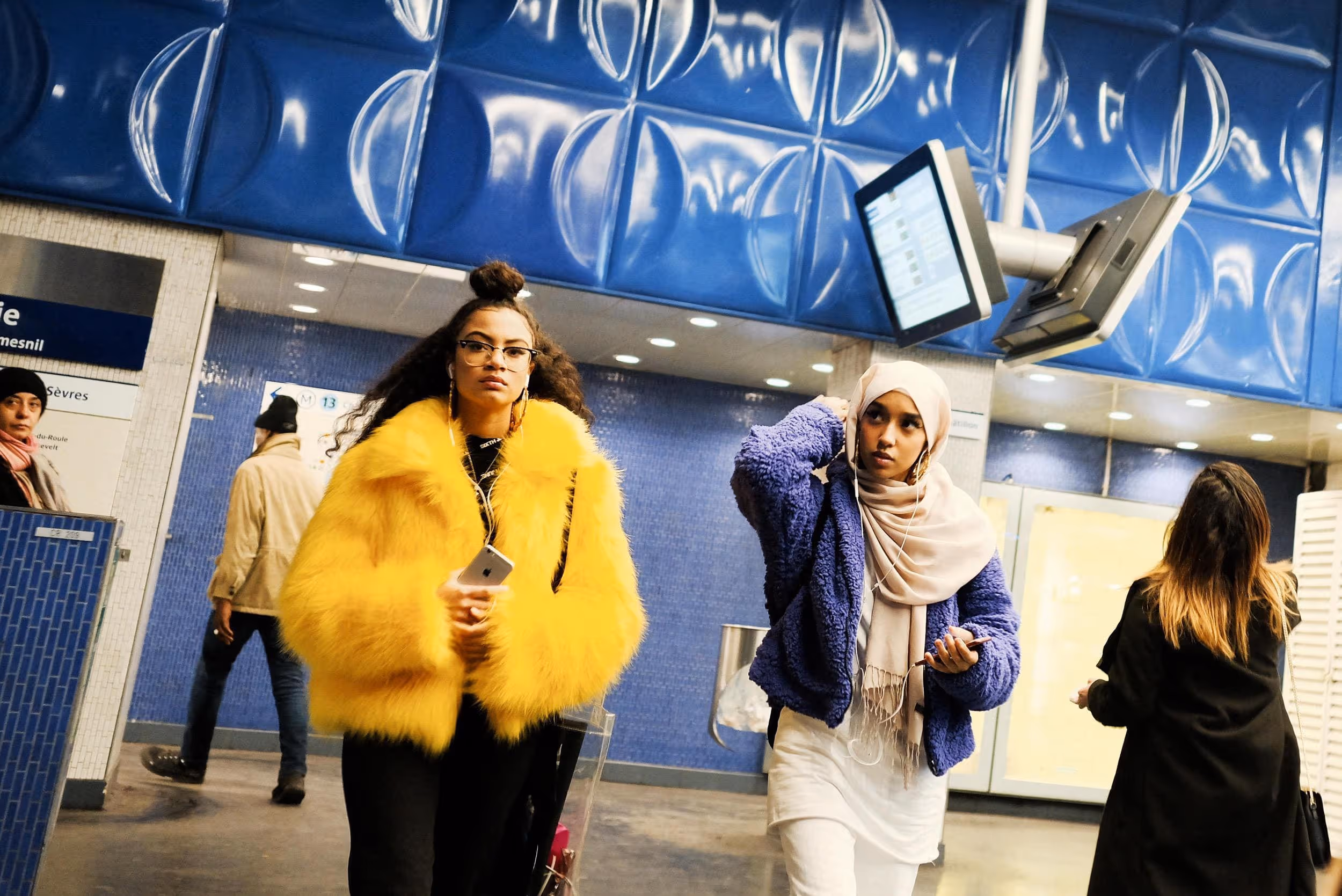 Deux jeunes femmes à la mode, l’une en manteau jaune et l’autre en violet, marchent dans une station de métro.
