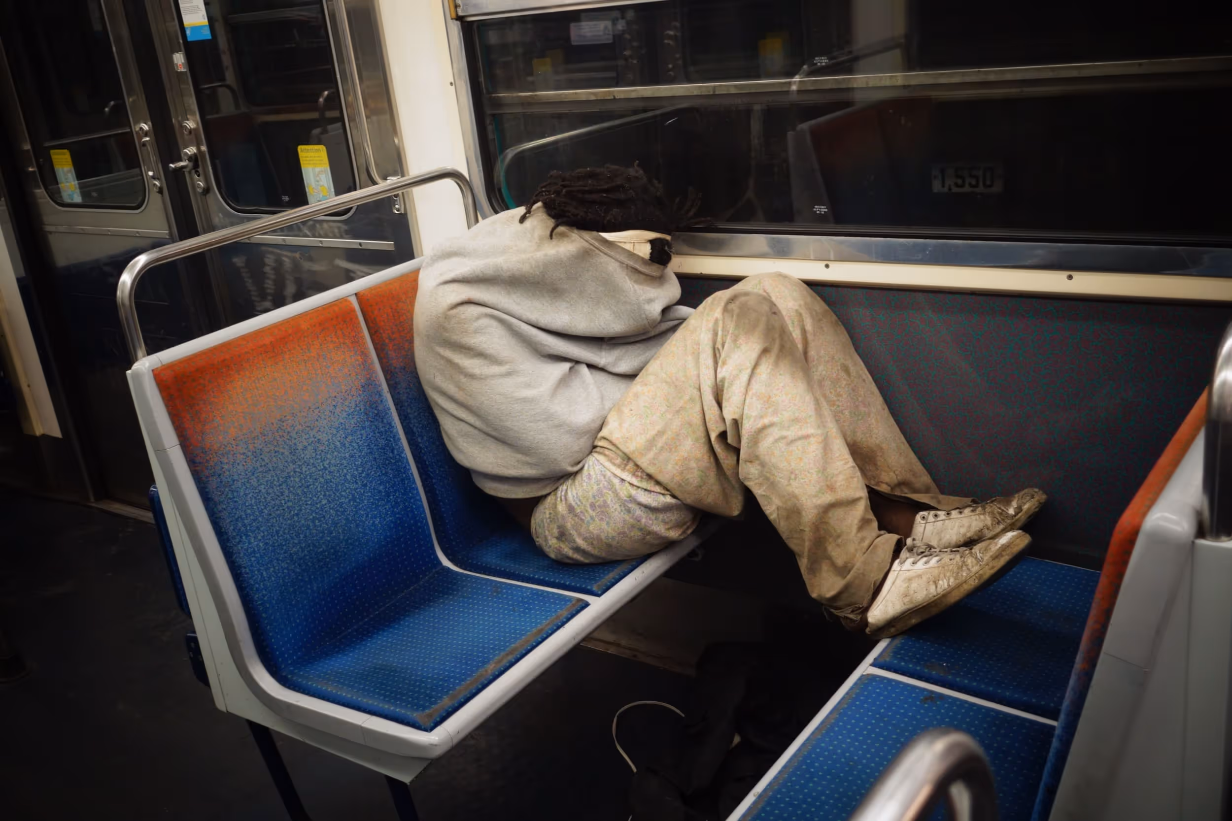 Personne recroquevillée sur une banquette de métro, habillée de vêtements sales et amples.