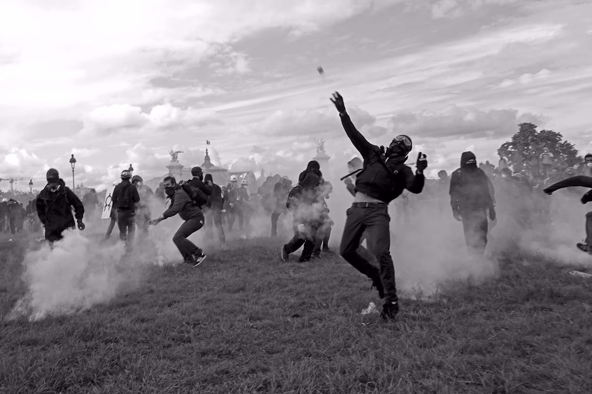 Manifestant masqué en train de jeter un projectile au milieu de la fumée, dans un champ, entouré d'autres protestataires.