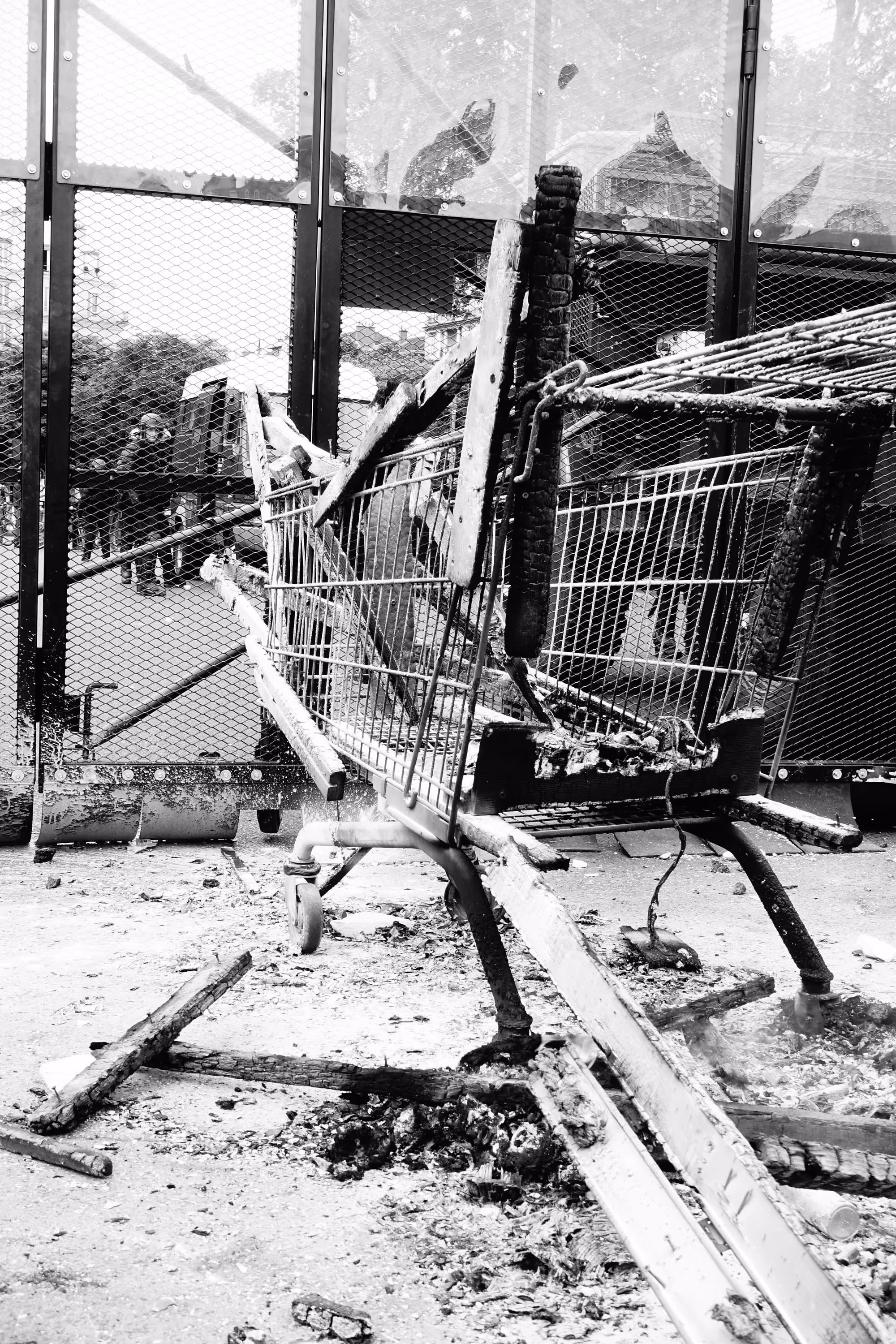 Caddie de supermarché brûlé formant une barricade devant des grilles métalliques lors d'une manifestation.