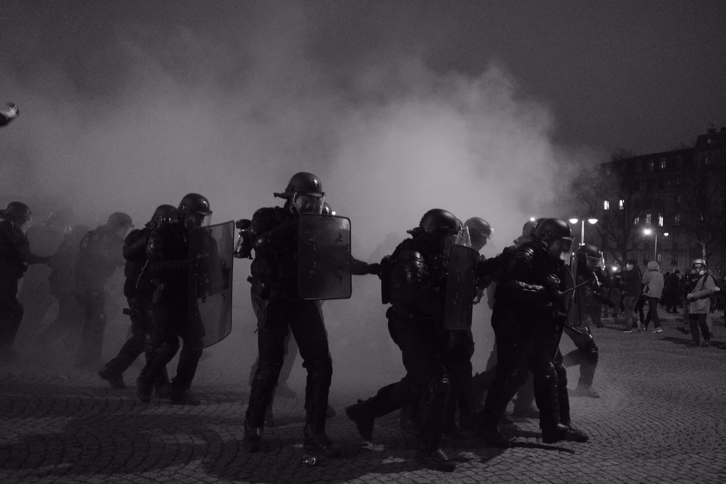 Ligne de policiers en tenue antiémeute avançant à travers un nuage de gaz lacrymogène.