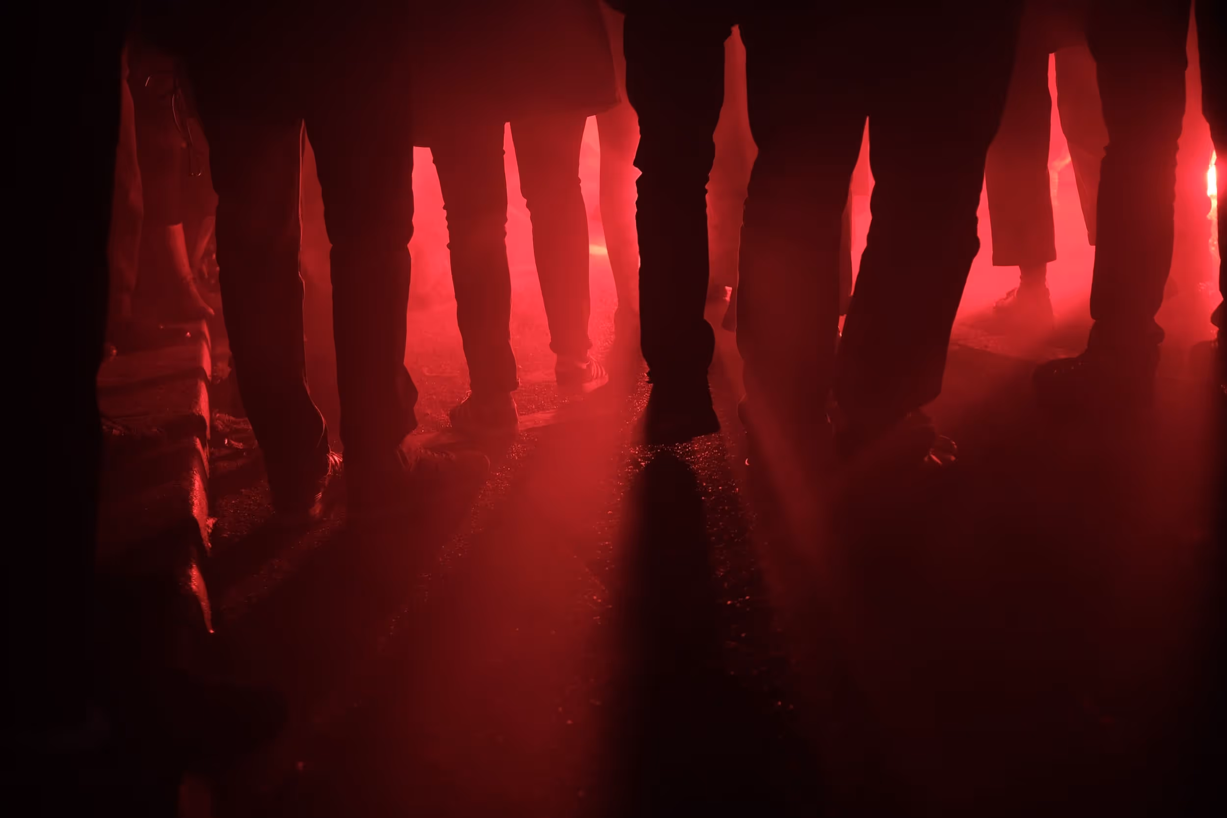 Silhouettes de jambes dans une rue enfumée, baignées d'une lumière rouge intense.