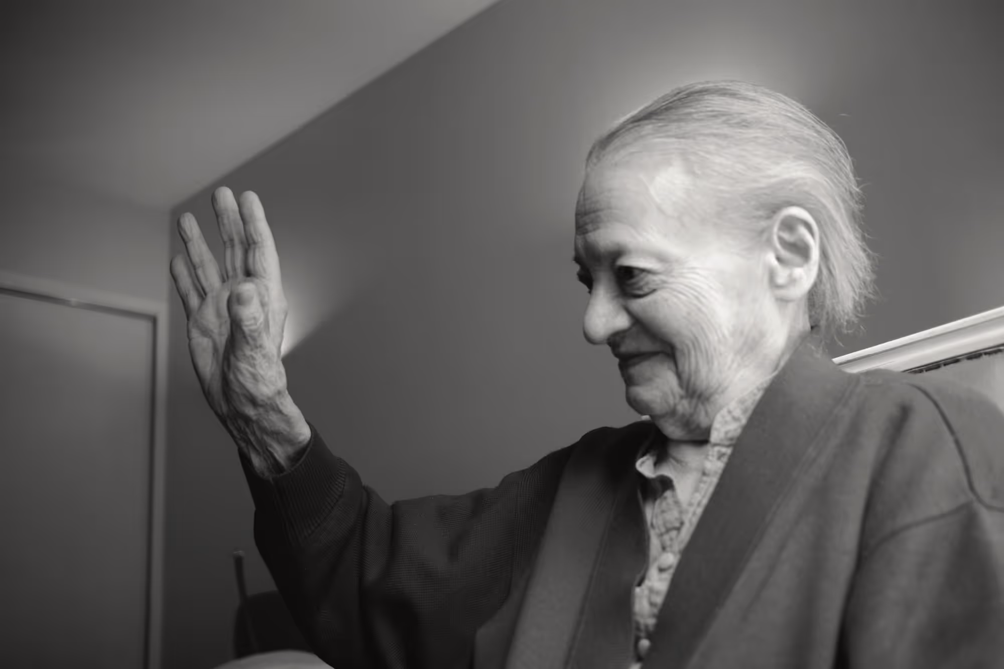 Femme âgée assise, levant la main en guise de salutation, sourire discret sur les lèvres.